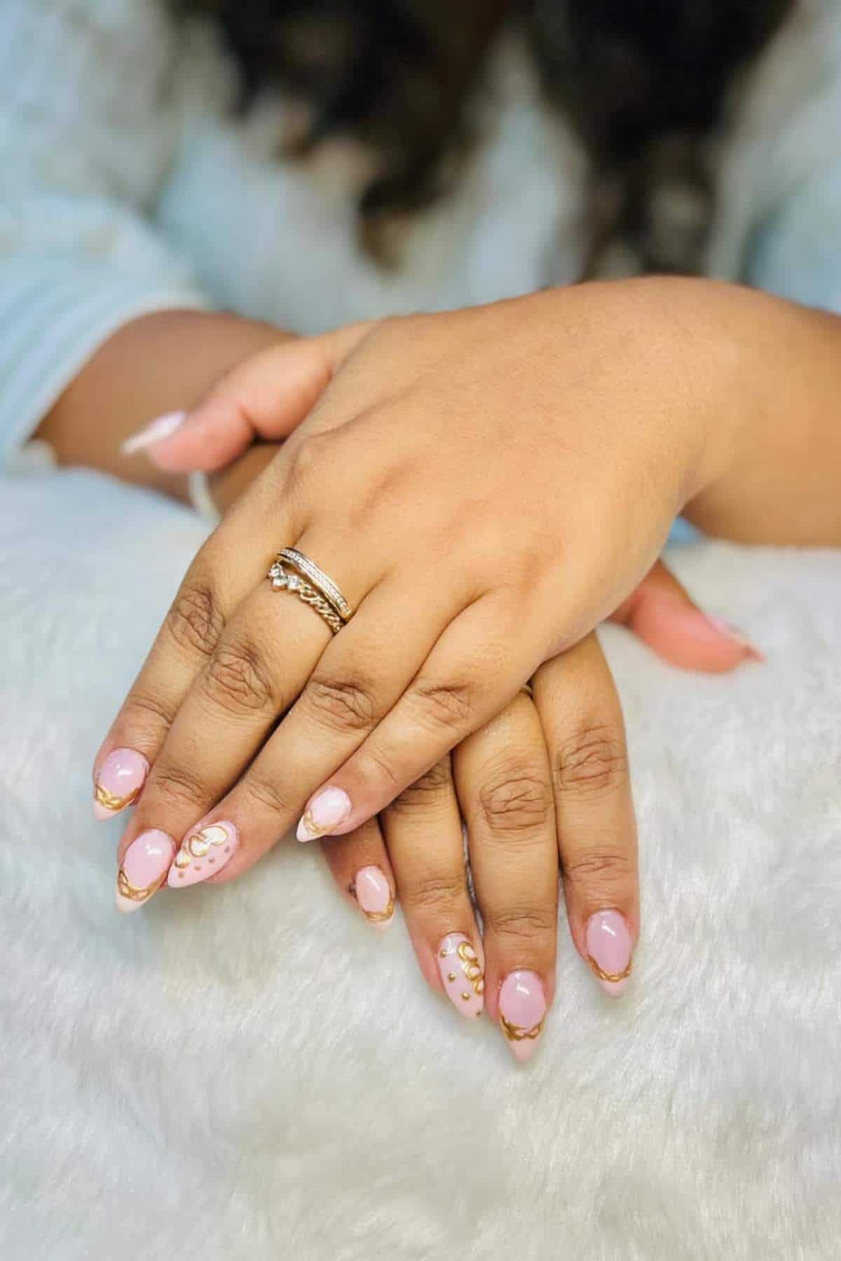 Hands resting on a white surface with pink and gold manicured nails, featuring decorative patterns and a gold ring on one finger.
