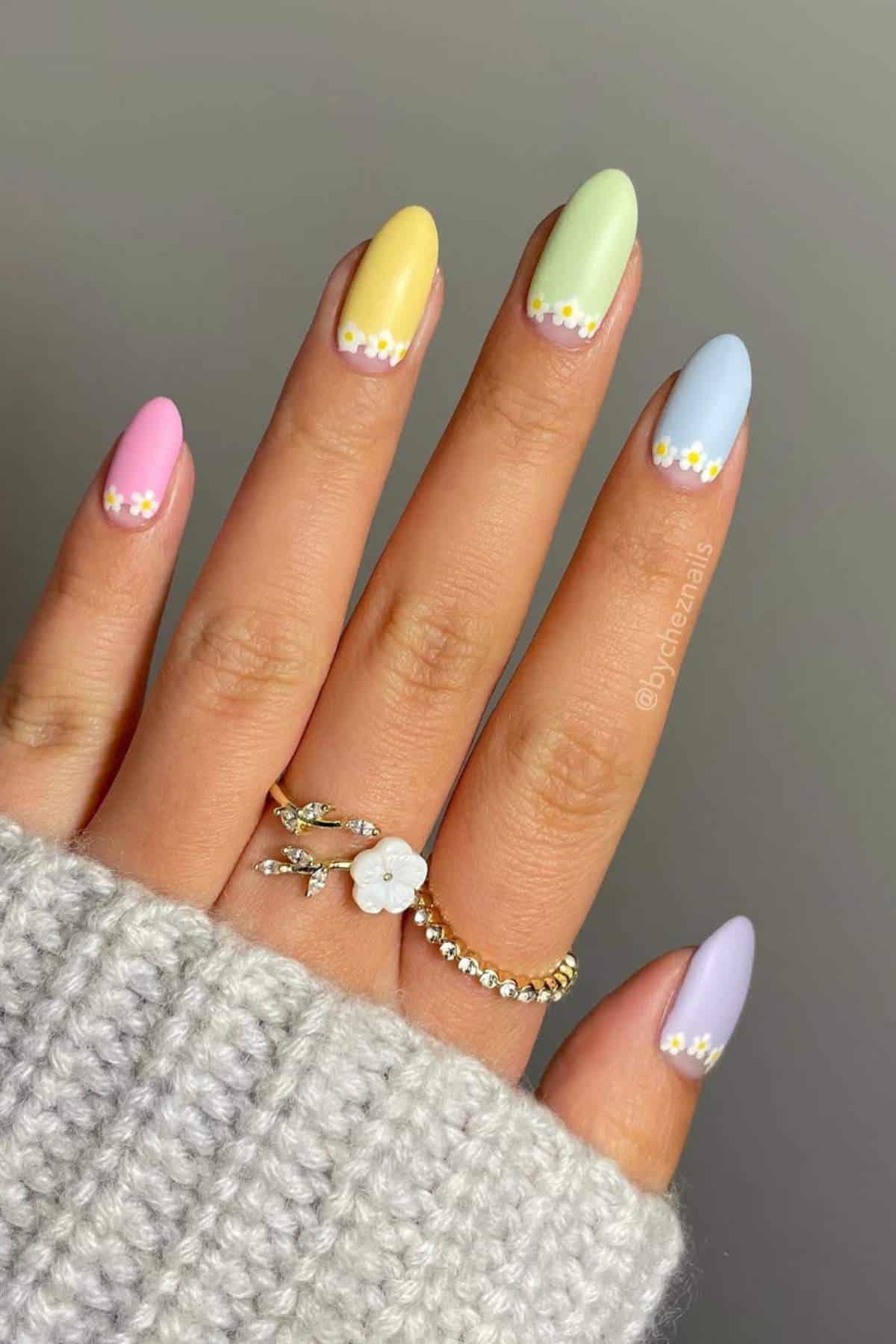 A hand with almond-shaped nails painted in pastel colors, each nail featuring small daisy designs near the tips. The person is wearing gold rings and a grey knit sleeve.