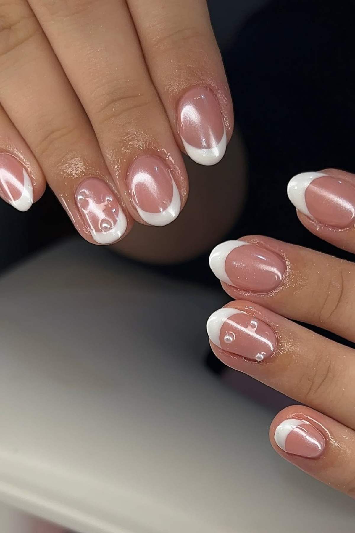 Hands with short, almond-shaped nails featuring a light pink base, white French tips, and subtle pearl-like accents on a few nails.