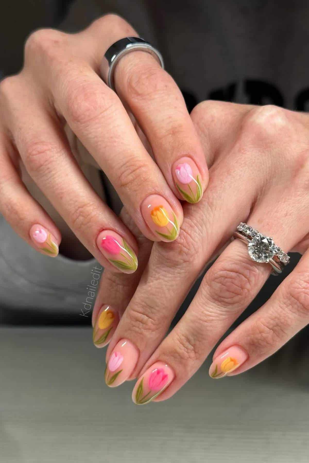 Close-up of hands with almond-shaped nails painted with pink and orange tulip designs and green leaves. Person wears silver rings, including a diamond engagement ring.