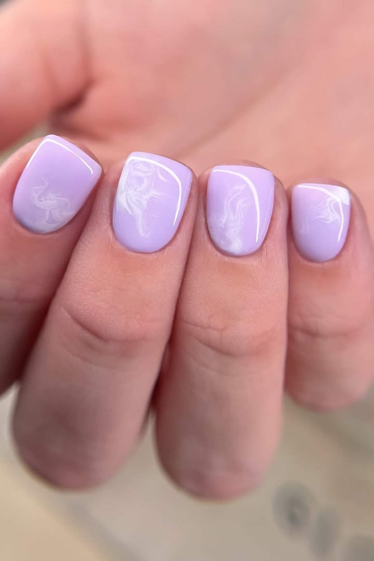 A hand with short, square-shaped nails painted light lavender with subtle white marble swirl designs on each nail.
