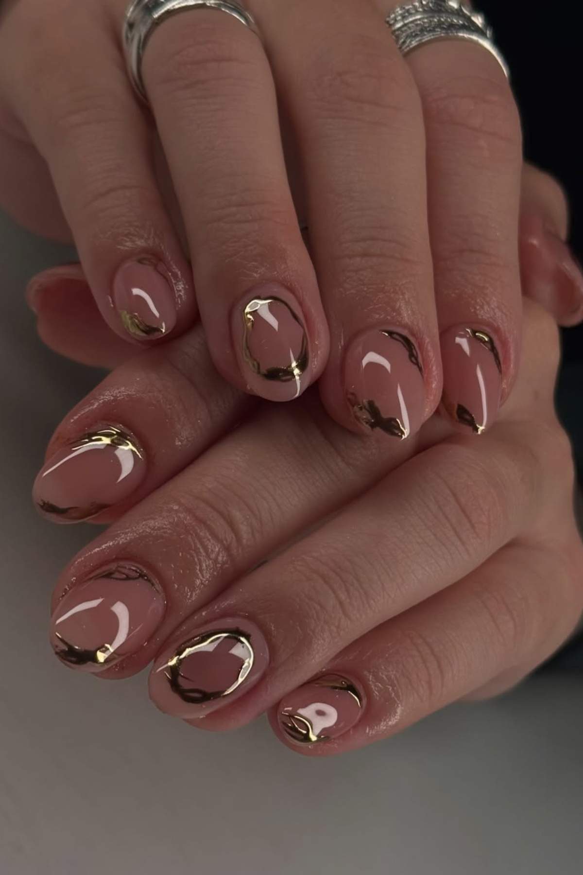 Close-up of hands with nude and gold abstract nail art designs; nails are short and oval-shaped, and fingers have silver rings.