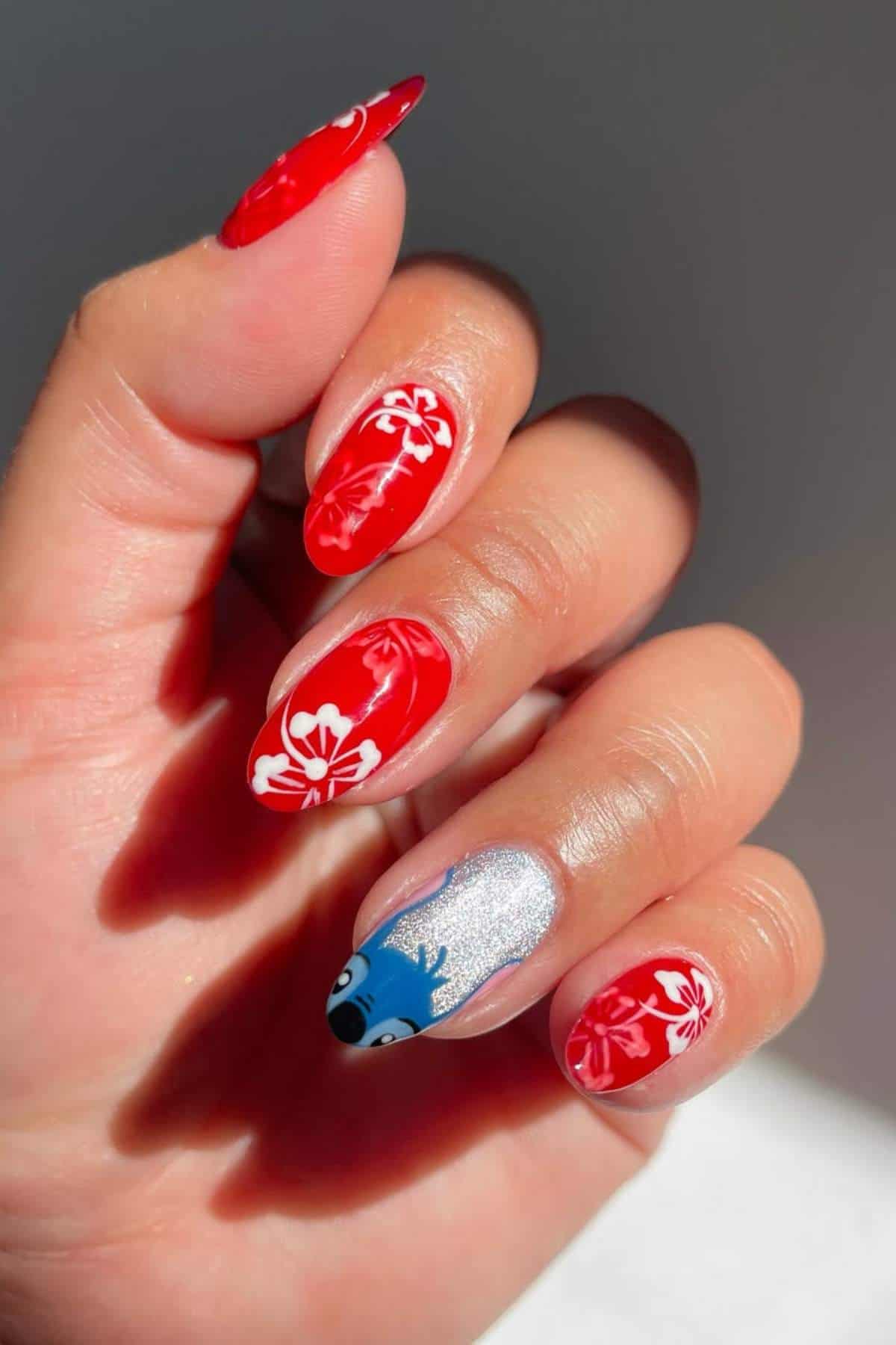A hand with manicured nails featuring red polish with white floral designs and one accent nail with silver polish and a blue cartoon character.