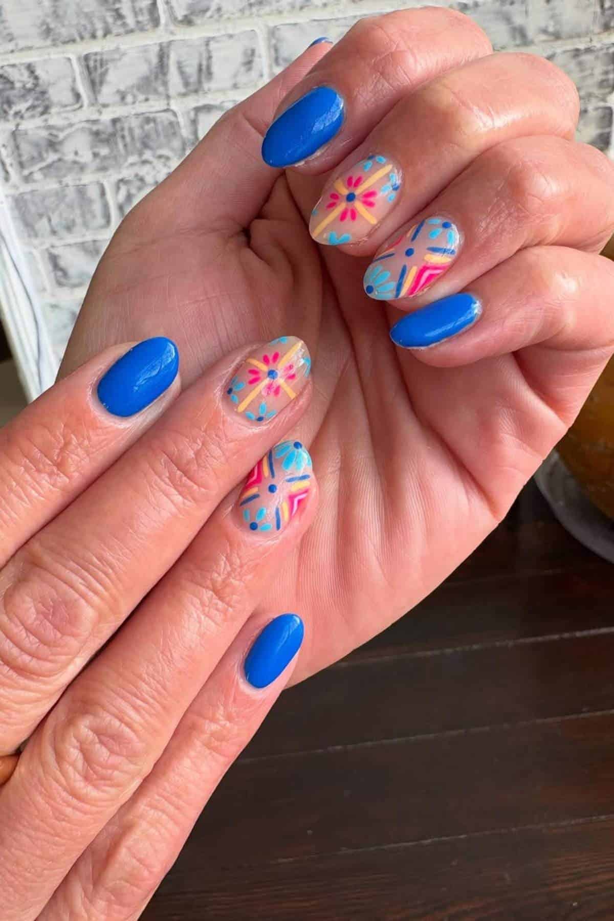 Hands with blue nail polish on some nails and colorful geometric and floral designs on others, set against a brick wall and dark wooden surface background.