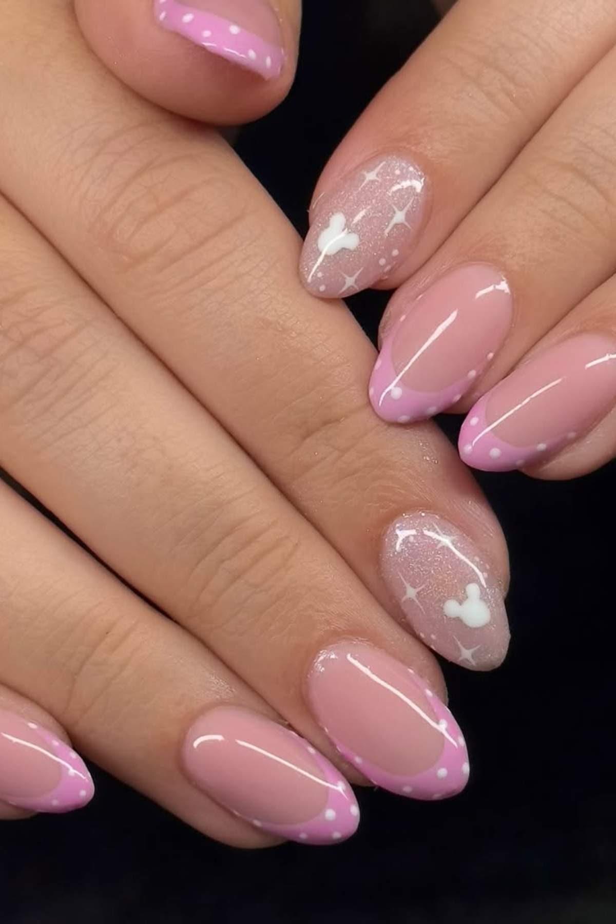 Hands with pink French tip nails, some featuring white polka dots and others decorated with white Mickey Mouse head shapes and small stars.