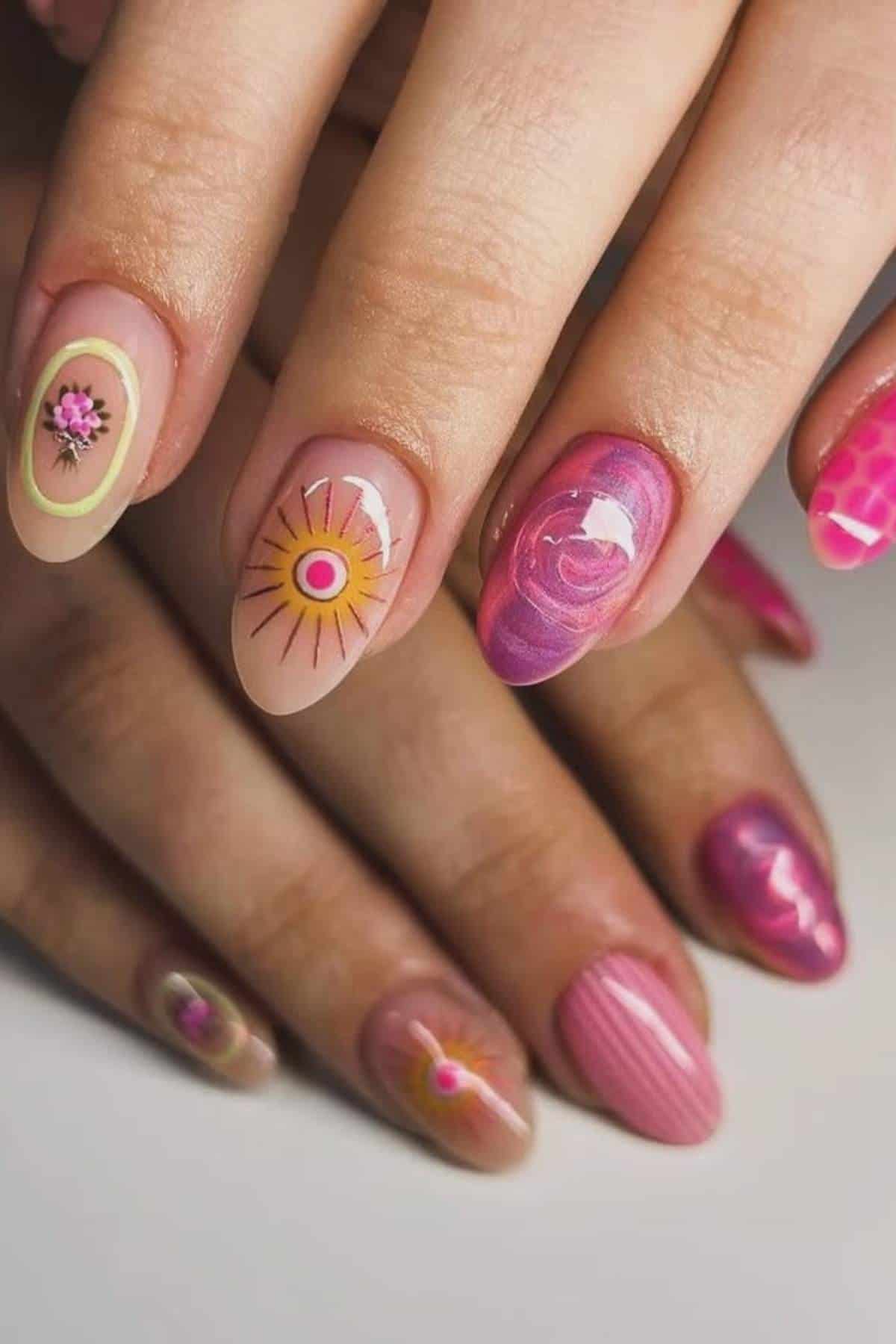Close-up of hands with manicured nails featuring pink, purple, and nude colors, some with abstract designs, flowers, and textured patterns.