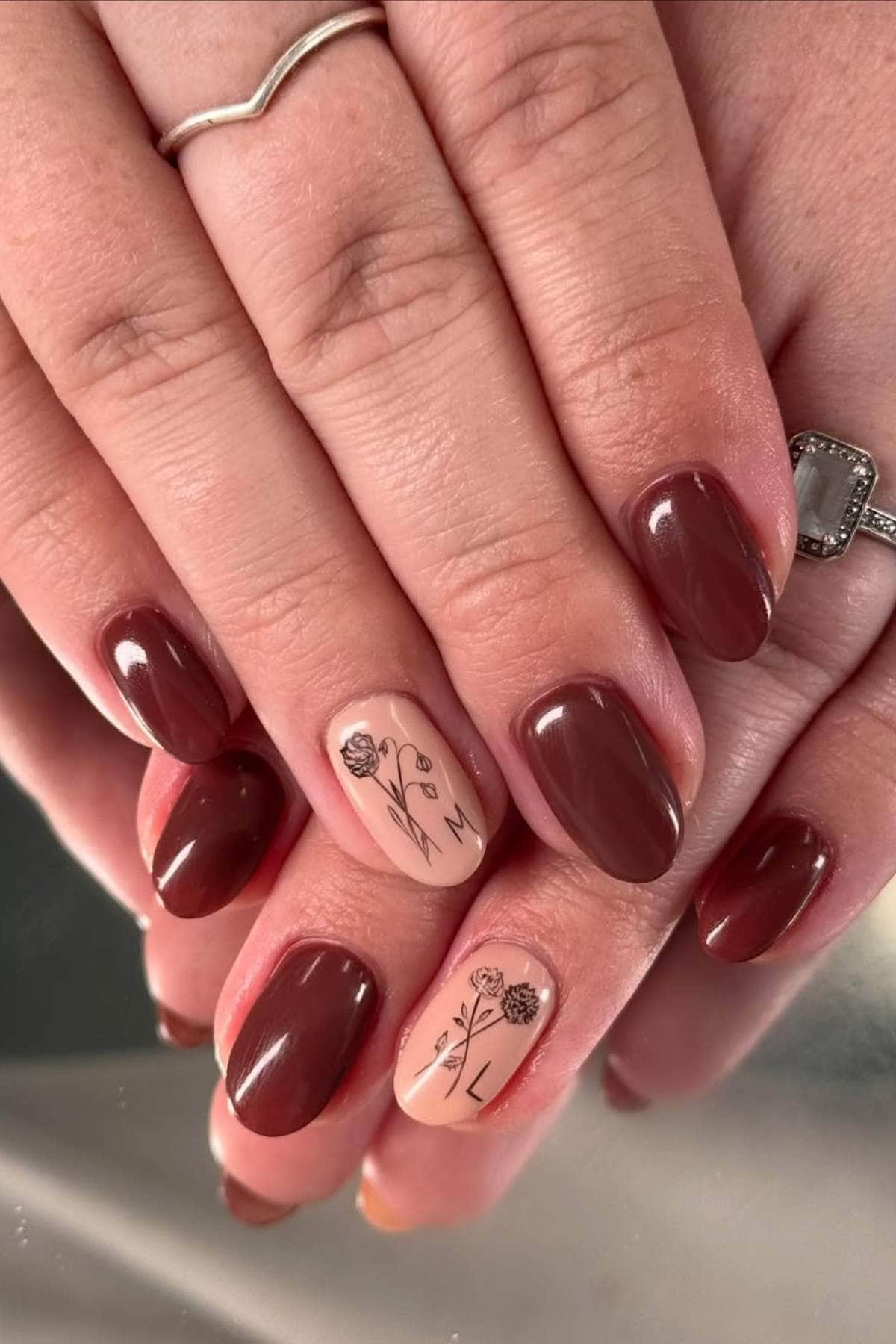 A hand with short, glossy brown nails; the ring and pinky fingers have floral line art and initials on a nude accent nail.
