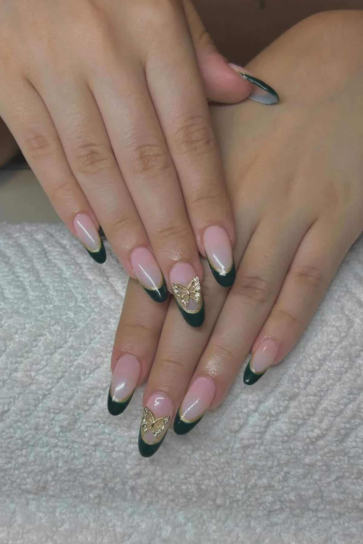 Hands with almond-shaped nails featuring dark green French tips, gold accents, and gold butterfly charms on the ring fingers, resting on a textured white surface.