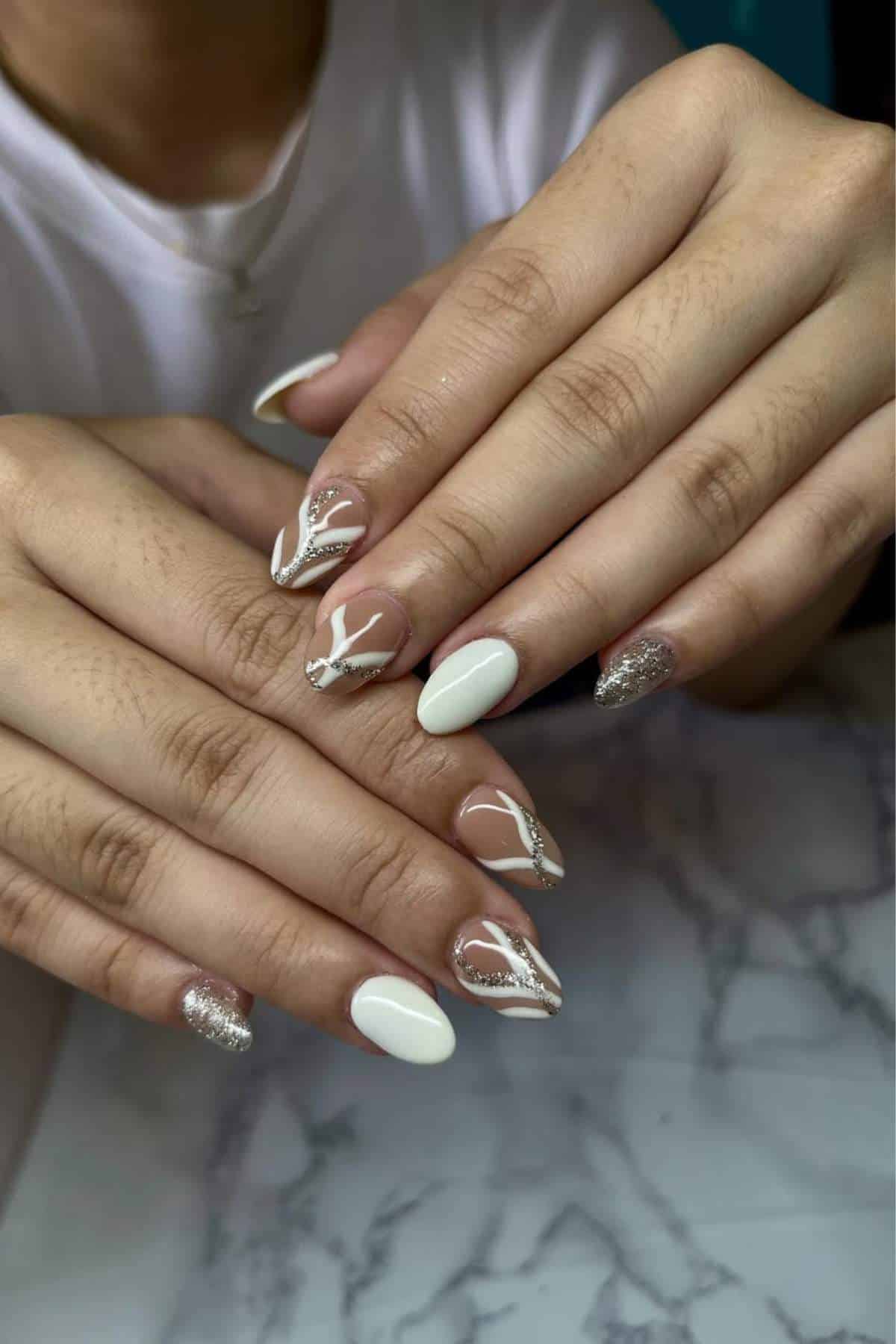 Close-up of hands with manicured nails featuring a white and nude base, silver glitter accents, and abstract white and silver line designs.