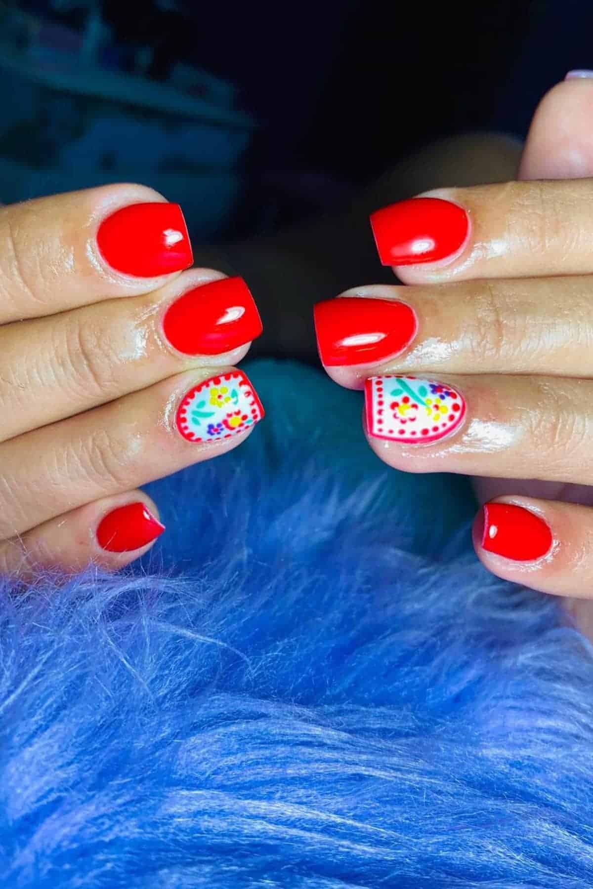 Hands with bright red square-shaped nails; ring fingernails feature a heart design with colorful dots and patterns on a white background.
