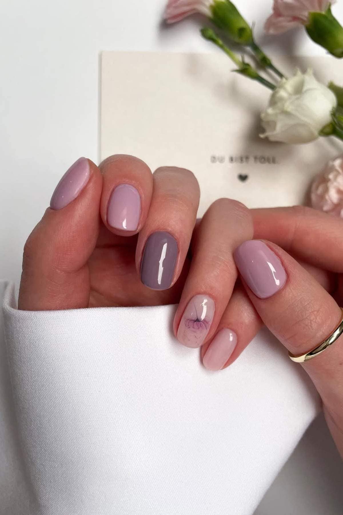 A hand with short, oval-shaped nails painted in shades of pink and mauve, one nail featuring a delicate floral accent; flowers and a white card appear in the background.