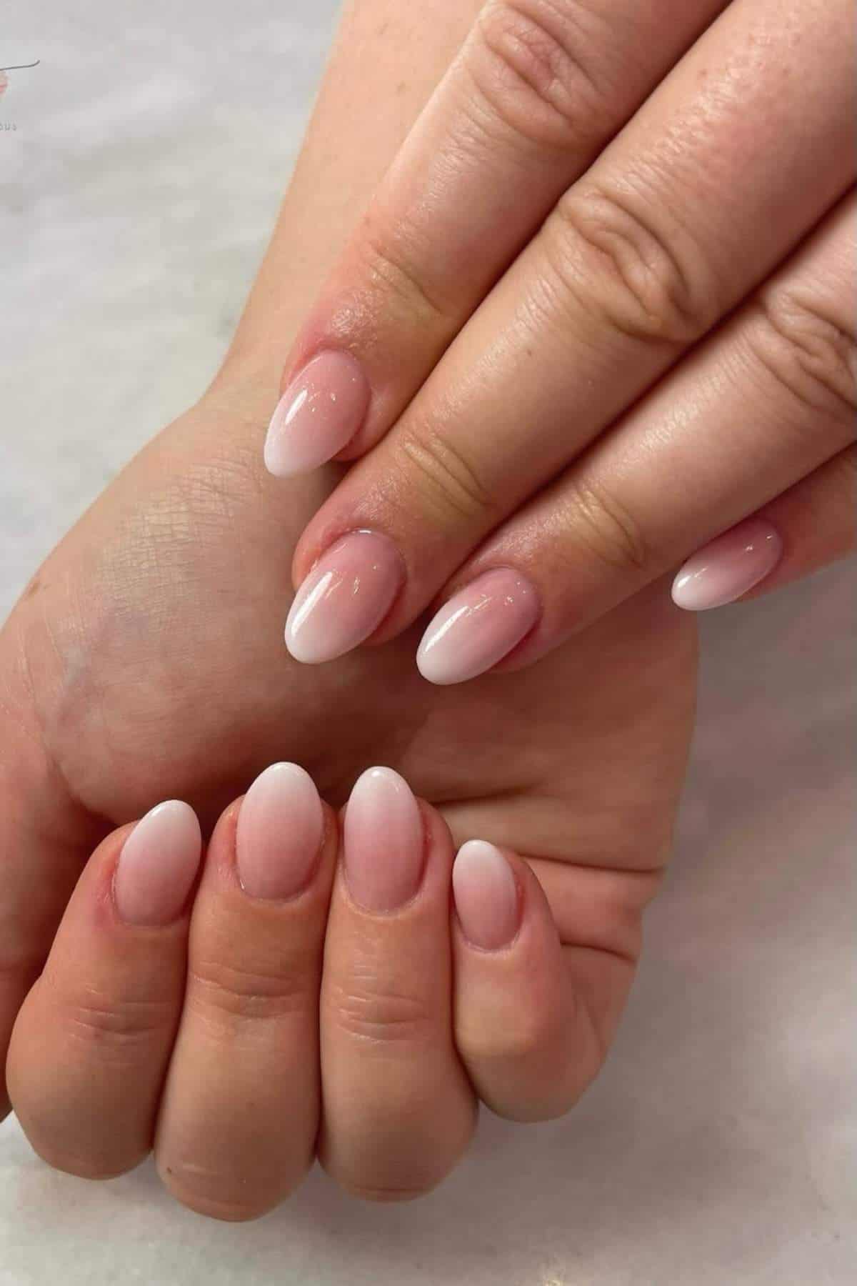 A hand displays almond-shaped nails with a light pink and white ombre manicure on a neutral background.