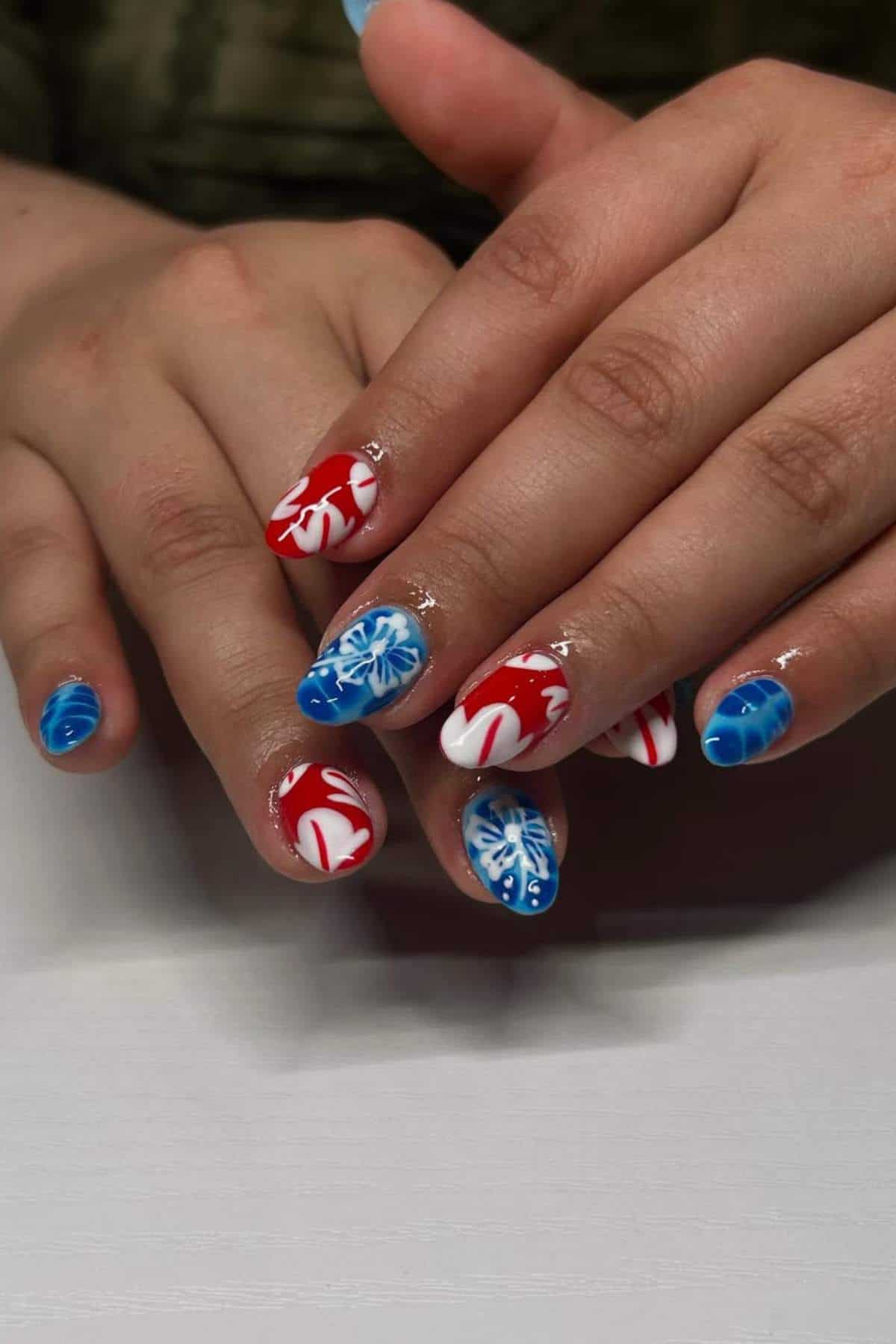 Hands with manicured nails featuring red, white, and blue designs; some nails have abstract patterns, others have floral or snowflake-like motifs.