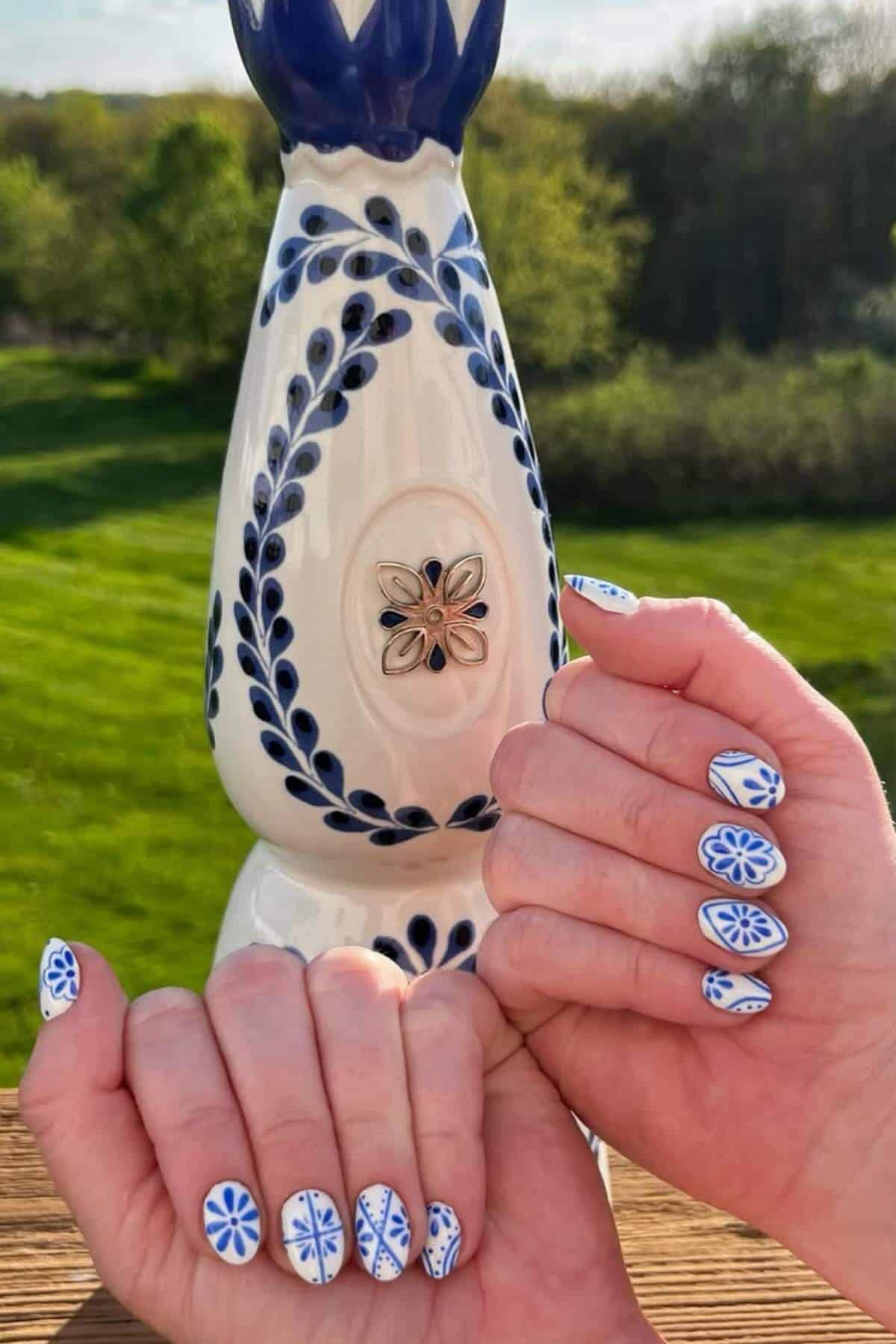 Two hands with blue and white floral nail art are held in front of a decorative ceramic vase featuring a similar blue and white pattern, with greenery in the background.
