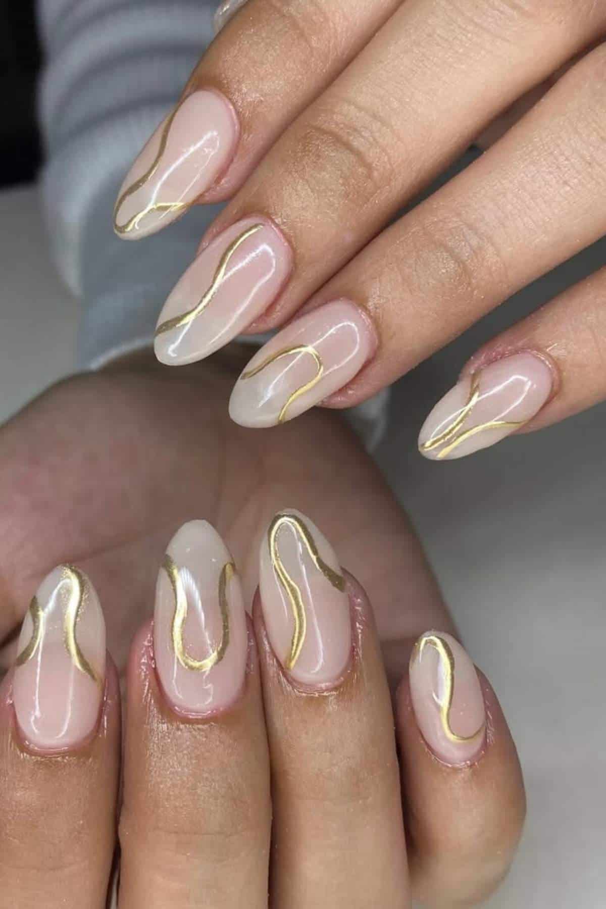 Close-up of hands with almond-shaped nails featuring a nude base and abstract gold line designs.