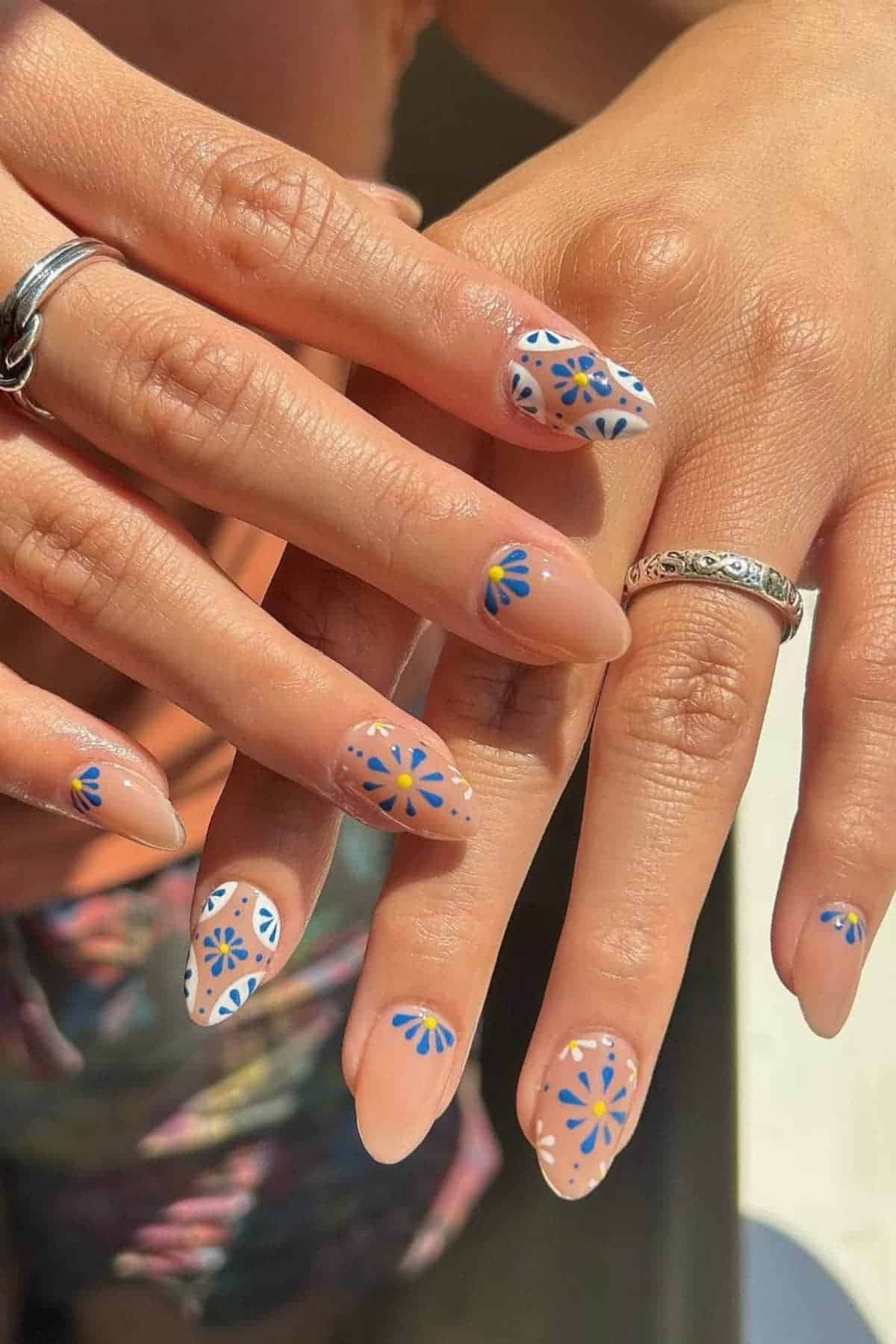 A close-up of hands with almond-shaped nails painted in nude, decorated with blue and yellow floral designs and white dotted patterns.