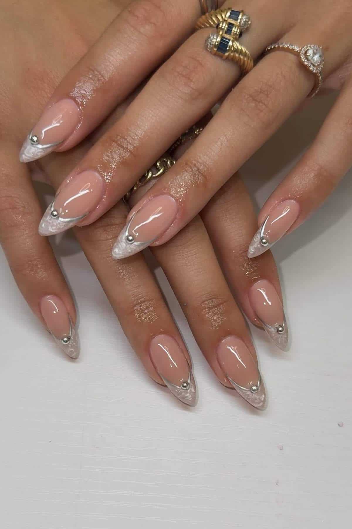 Hands with almond-shaped nails featuring a nude base, clear tips, and small silver studs. The person is wearing several gold and silver rings.