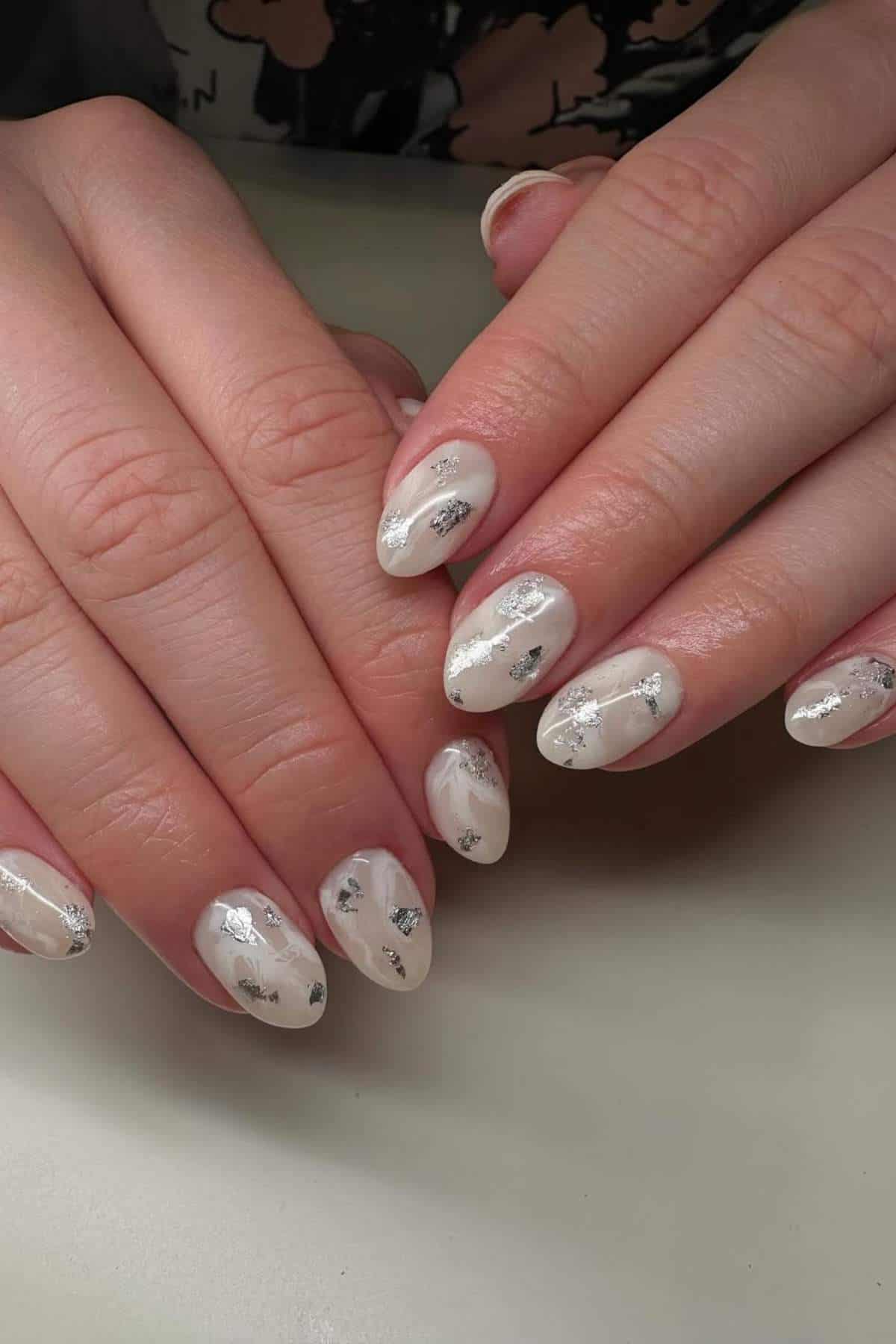 Hands with almond-shaped nails painted white and decorated with irregular silver foil accents.