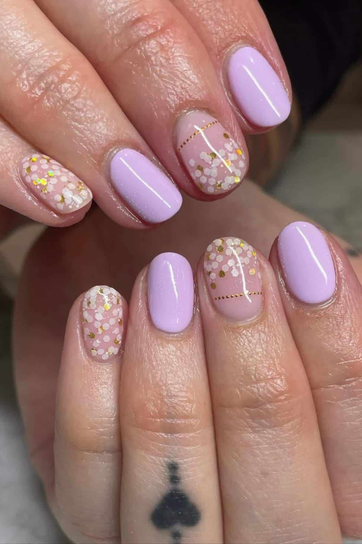 Hands with light purple and transparent nails featuring white, gold, and pink floral dot designs. An ace of spades tattoo is visible on the finger.