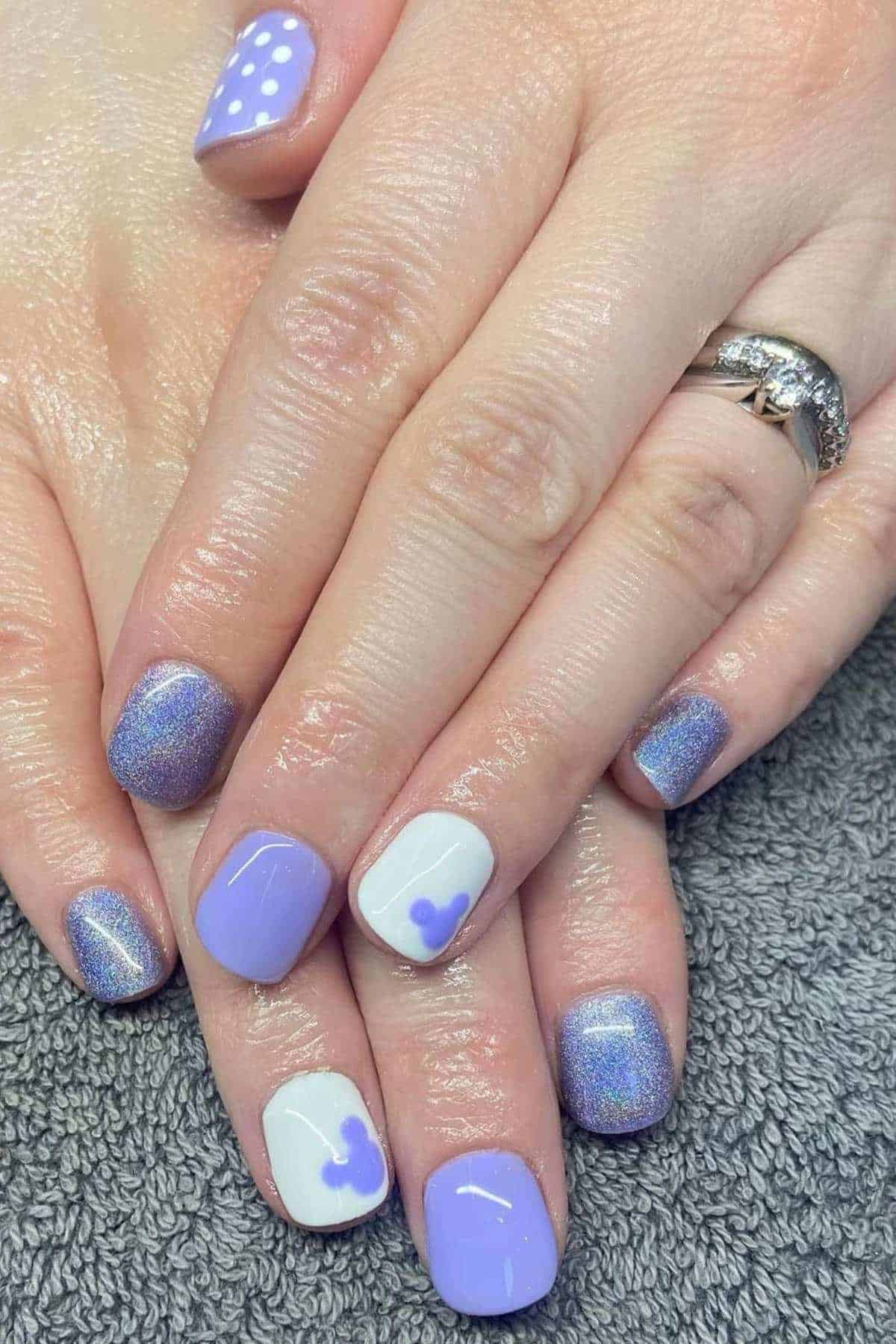 Hands with short nails featuring a purple and white manicure; some nails have shimmer, others have heart and polka dot designs.