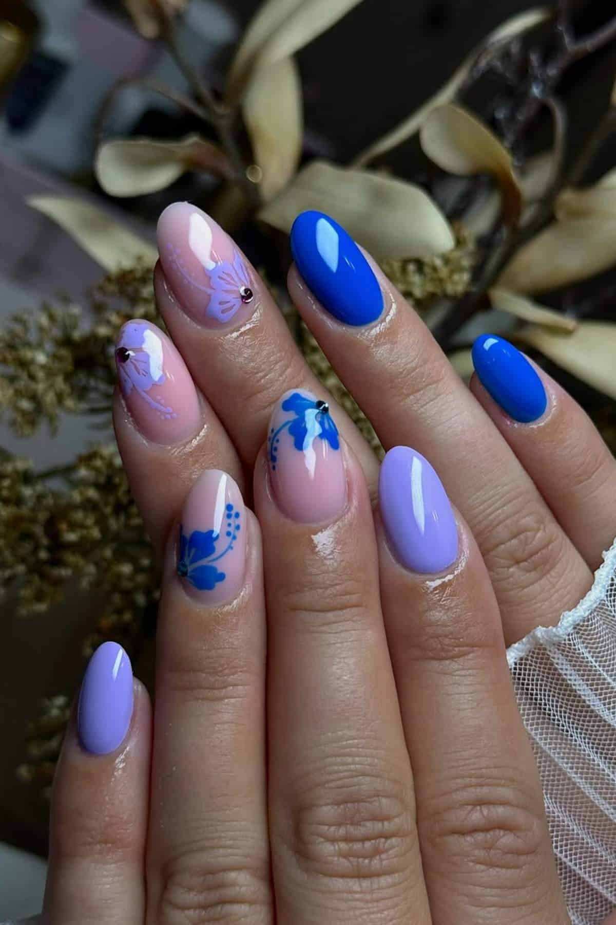 A hand with manicured nails painted in blue, lavender, and nude shades, featuring butterfly nail art designs on some fingernails. Dried plants are in the background.