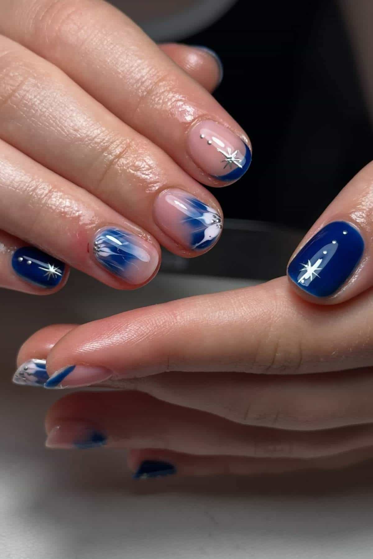 Close-up of hands with manicured nails featuring blue polish, white star designs, and blue and white abstract patterns on a few nails.