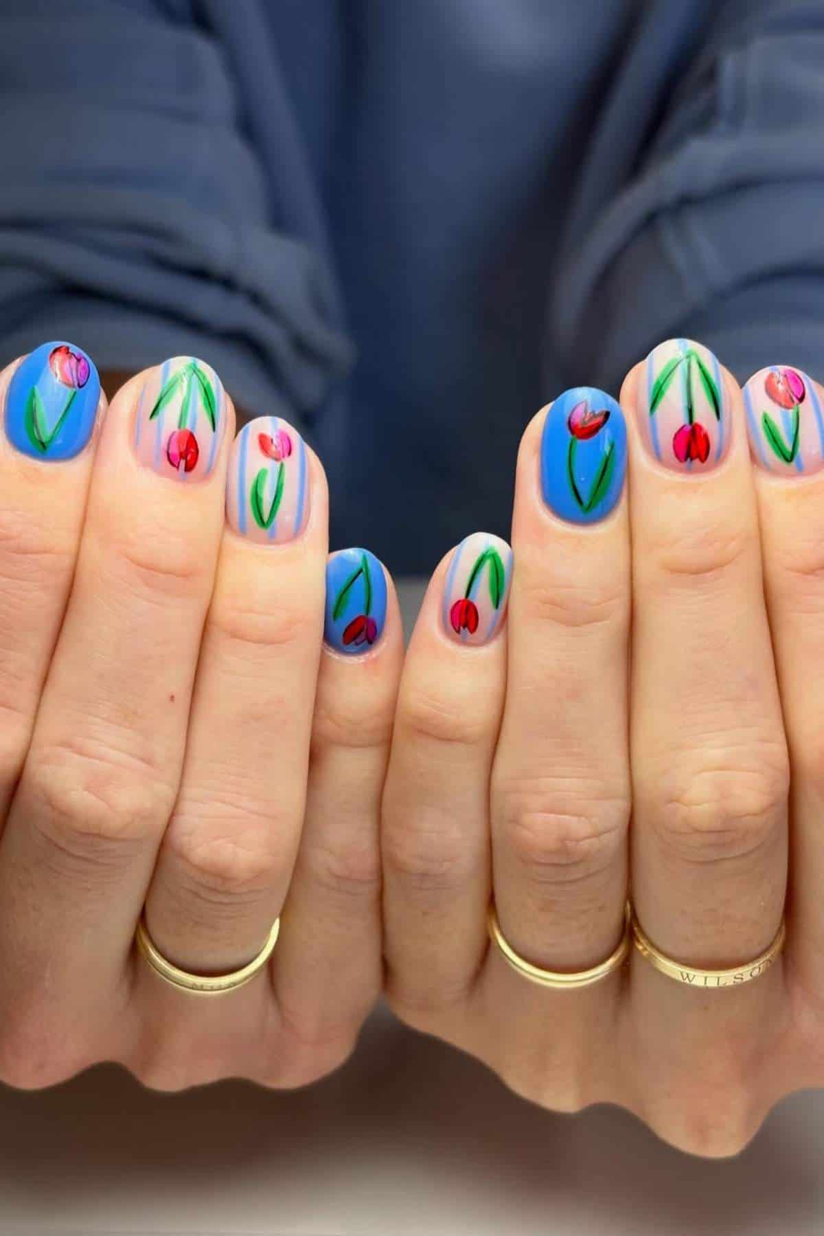 Close-up of hands with blue and nude nails featuring red tulip and green stem nail art; person is wearing gold rings.