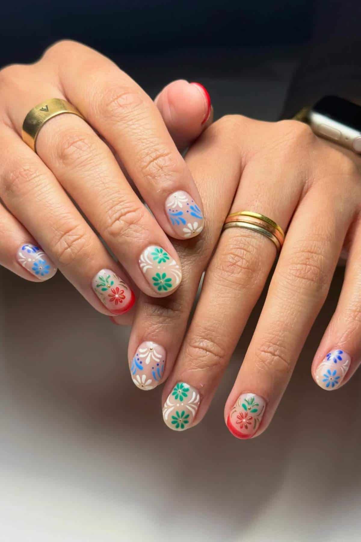 Close-up of hands with gold rings and a smartwatch, displaying manicure with colorful floral designs in blue, green, and red on short, rounded nails.