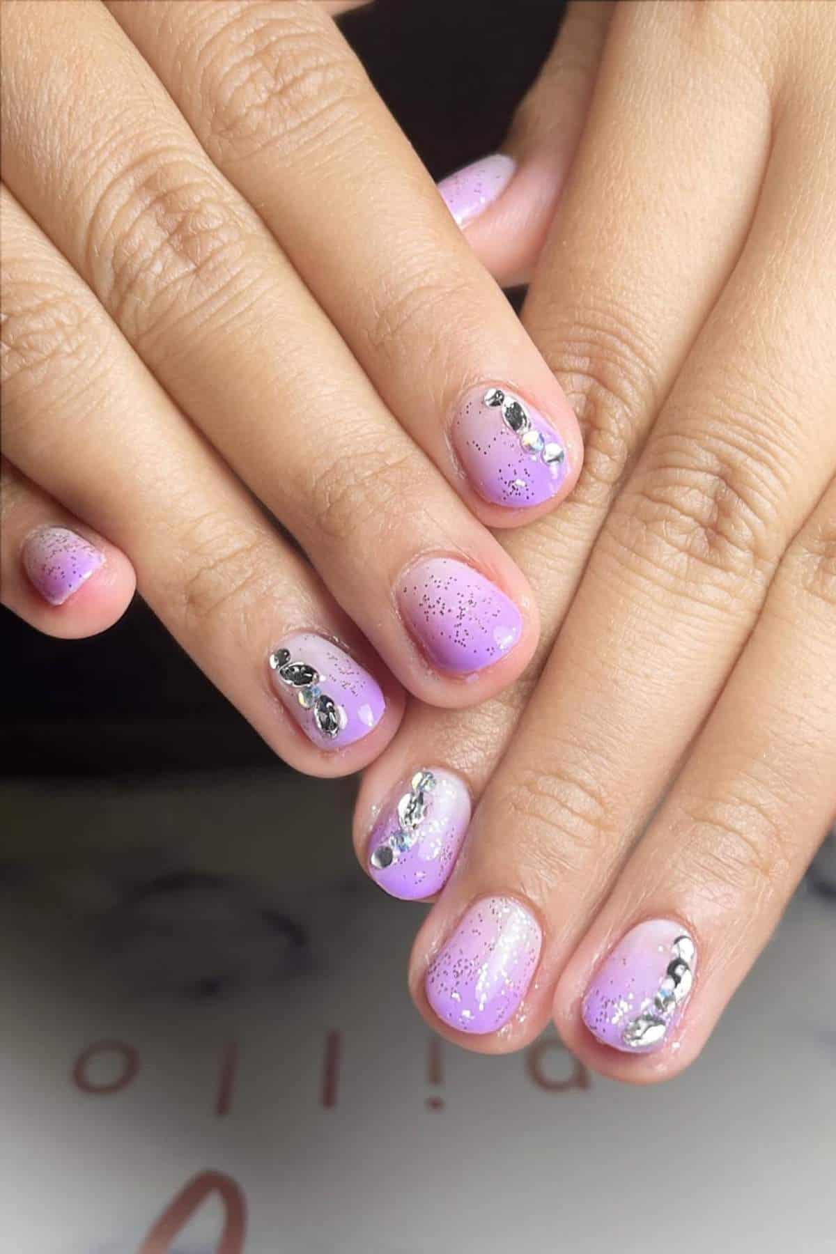 Hands with short nails painted in a gradient of light purple and pink, accented with silver glitter and small metallic decorations on some nails.