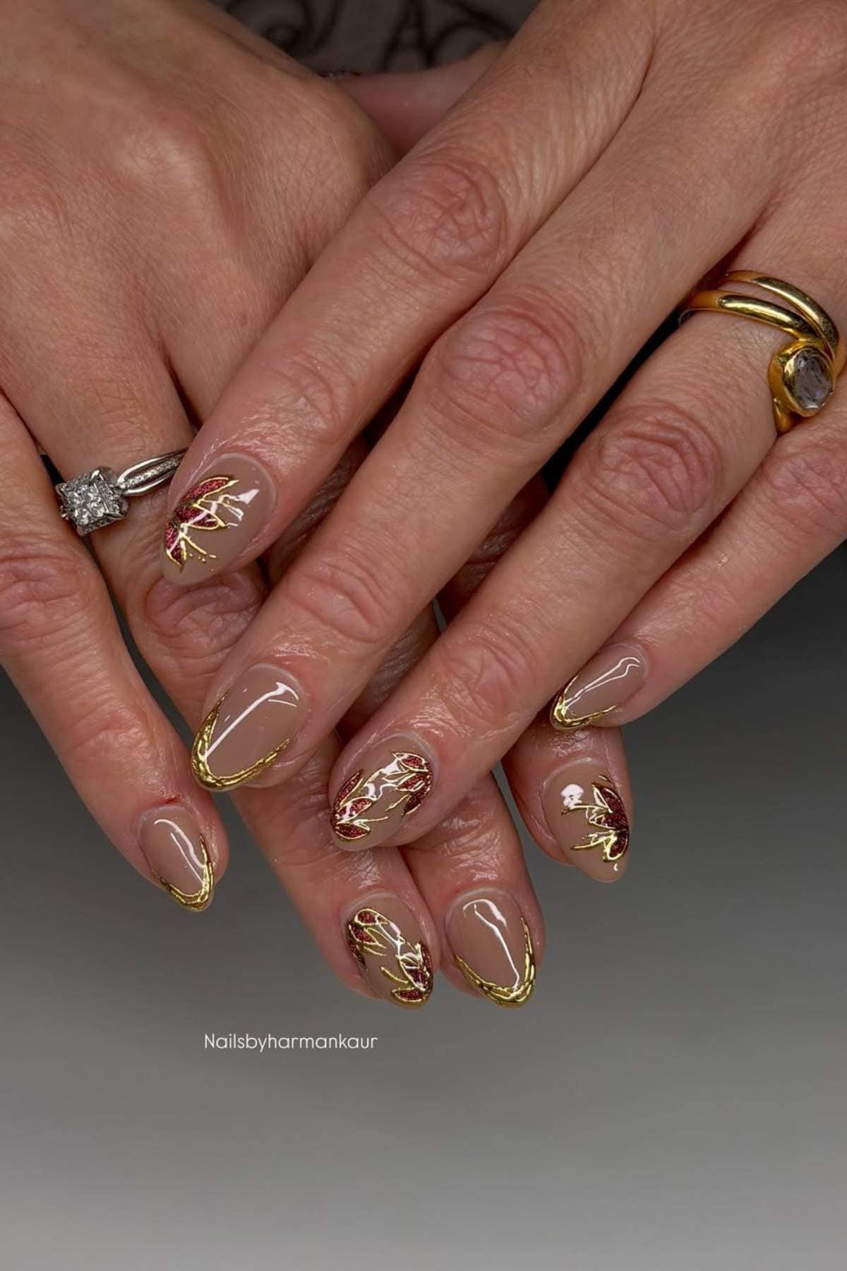 Close-up of hands with almond-shaped nails featuring nude polish, gold accents, and red marble designs; person is wearing two rings, including one with a large gemstone.