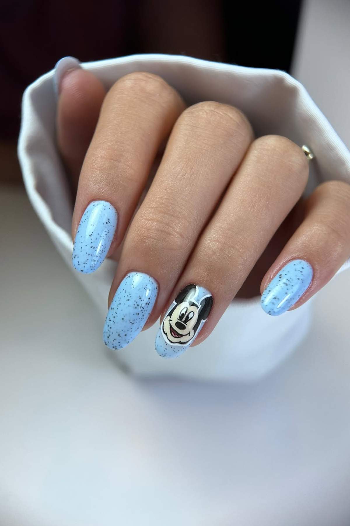 A hand with long, oval-shaped nails painted light blue with black speckles; one nail features a cartoon character's face.