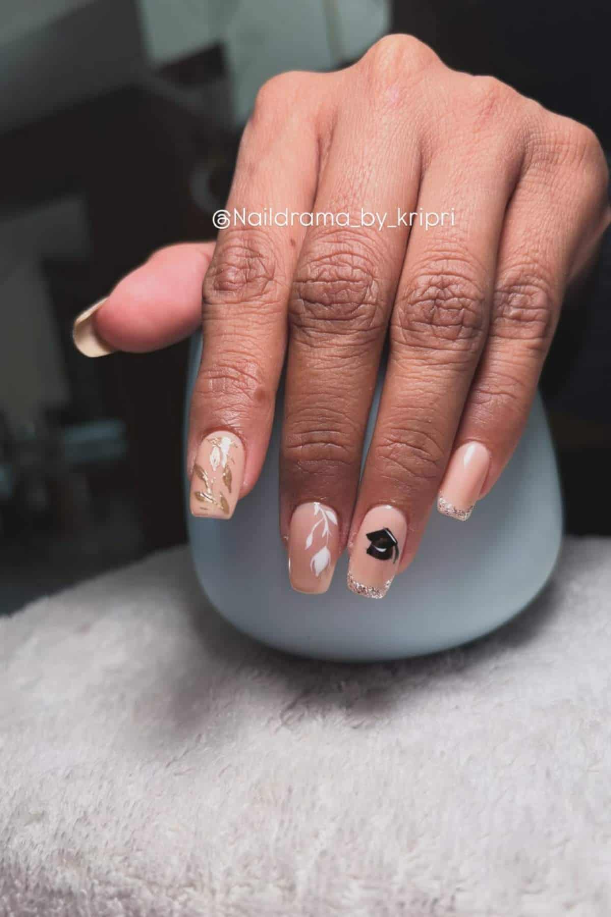 A hand with nude and beige ombre nails featuring black and gold leaf accents rests on a light gray surface.