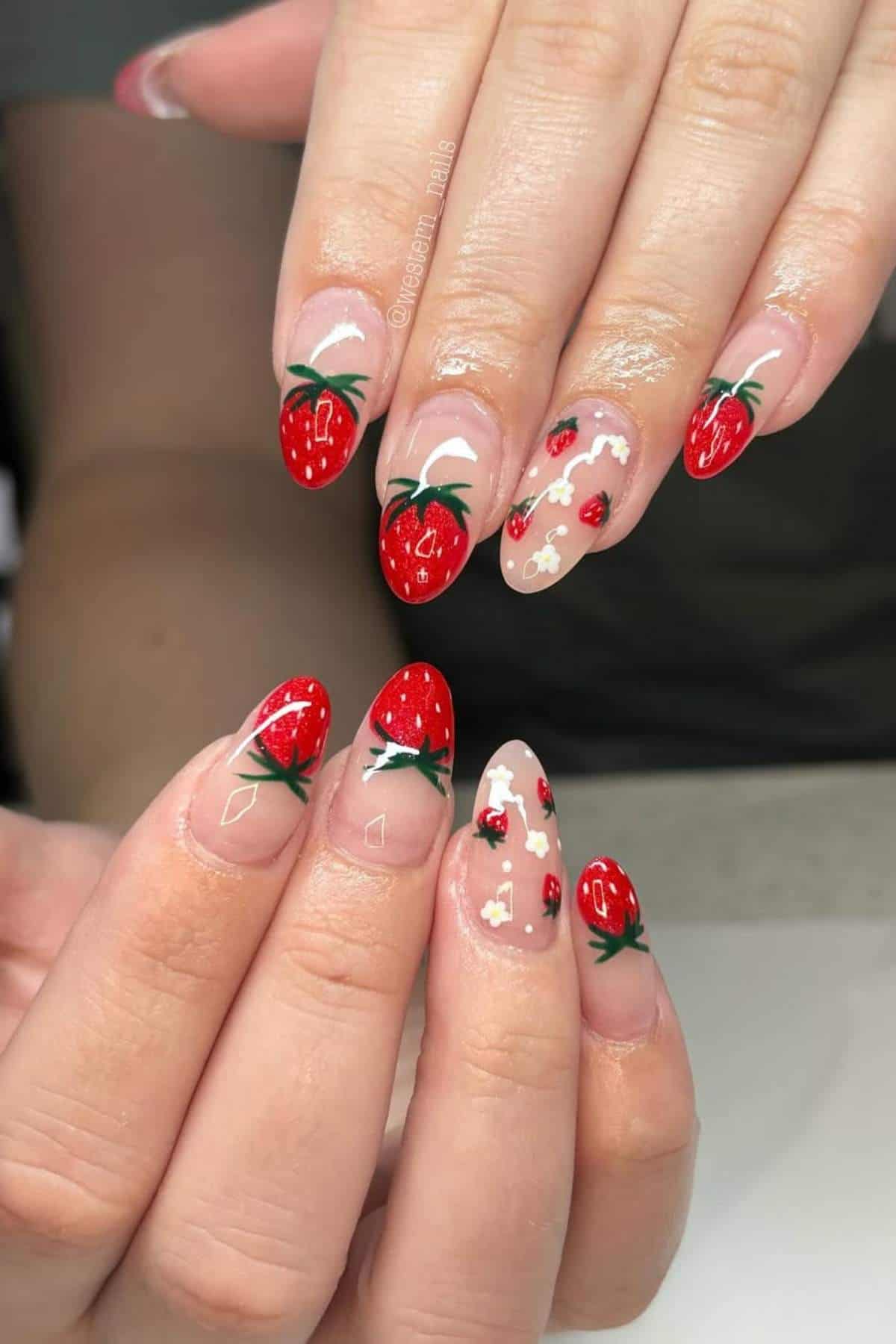 Hands with almond-shaped nails decorated with red strawberry and white flower nail art on a nude base.