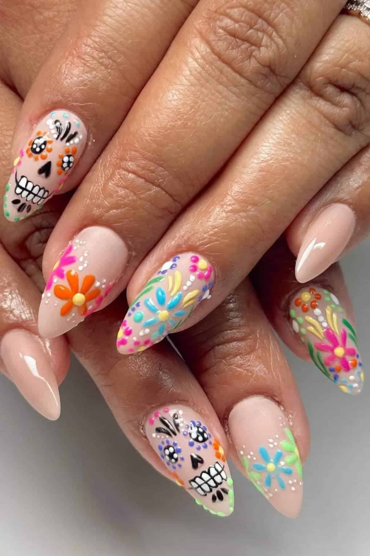 A hand with almond-shaped nails features colorful floral designs and Day of the Dead skull art on a nude base.