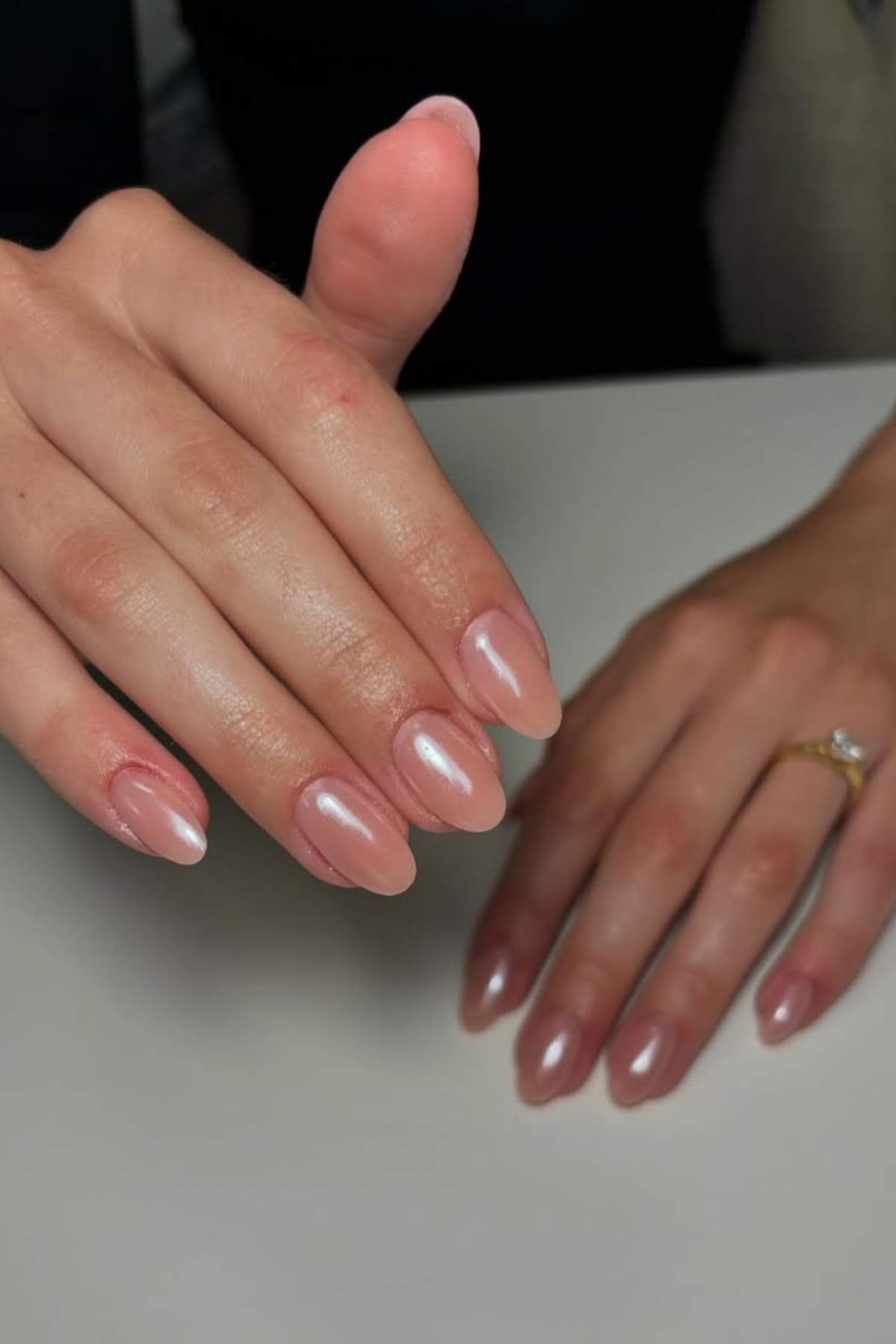 A hand with almond-shaped nails painted in a glossy nude color rests on a white surface; another hand wears a gold ring with a clear stone.