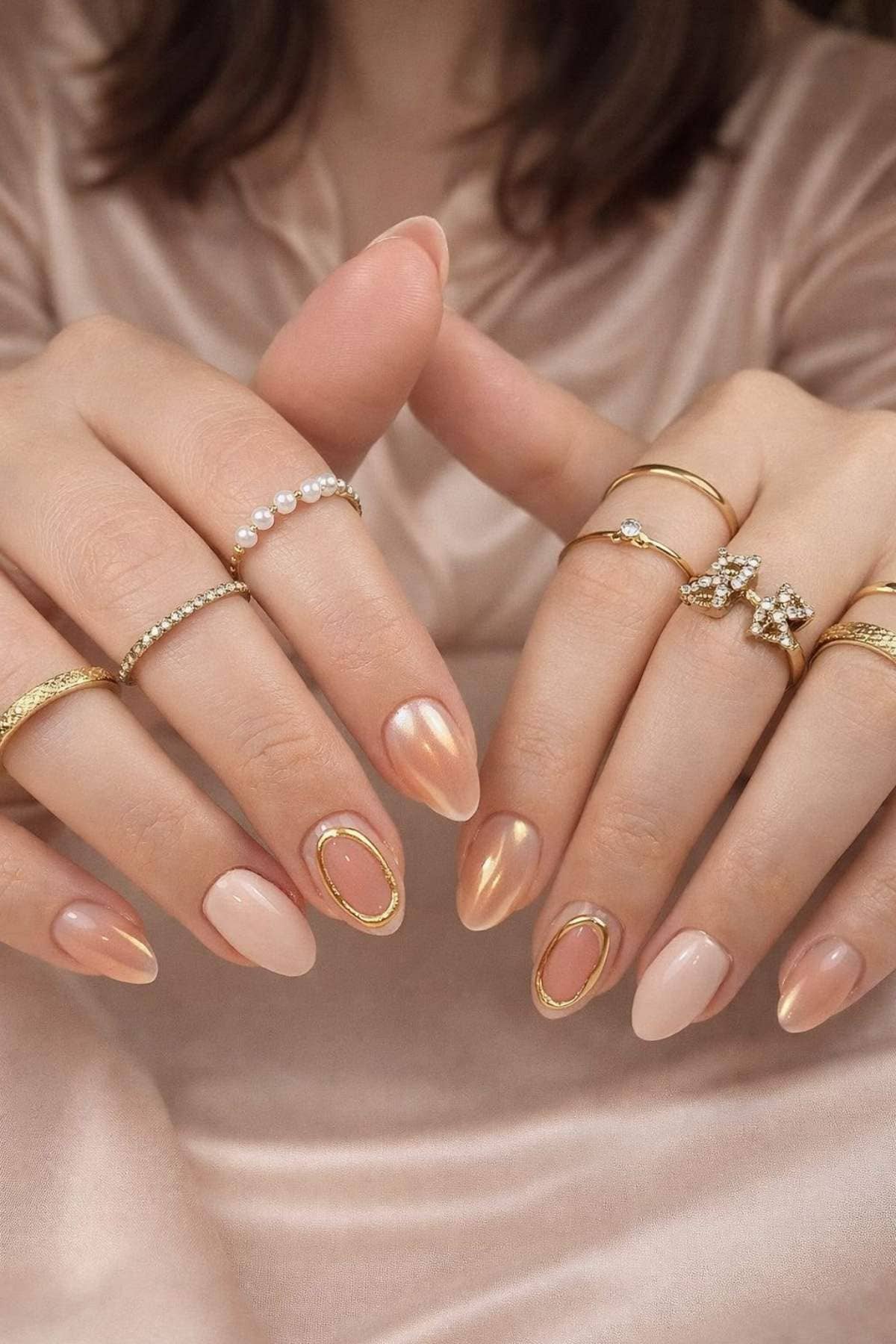 Hands with almond-shaped, nude and gold accent nail art. Multiple gold and pearl rings are worn on each finger. Person is wearing a light beige satin outfit.