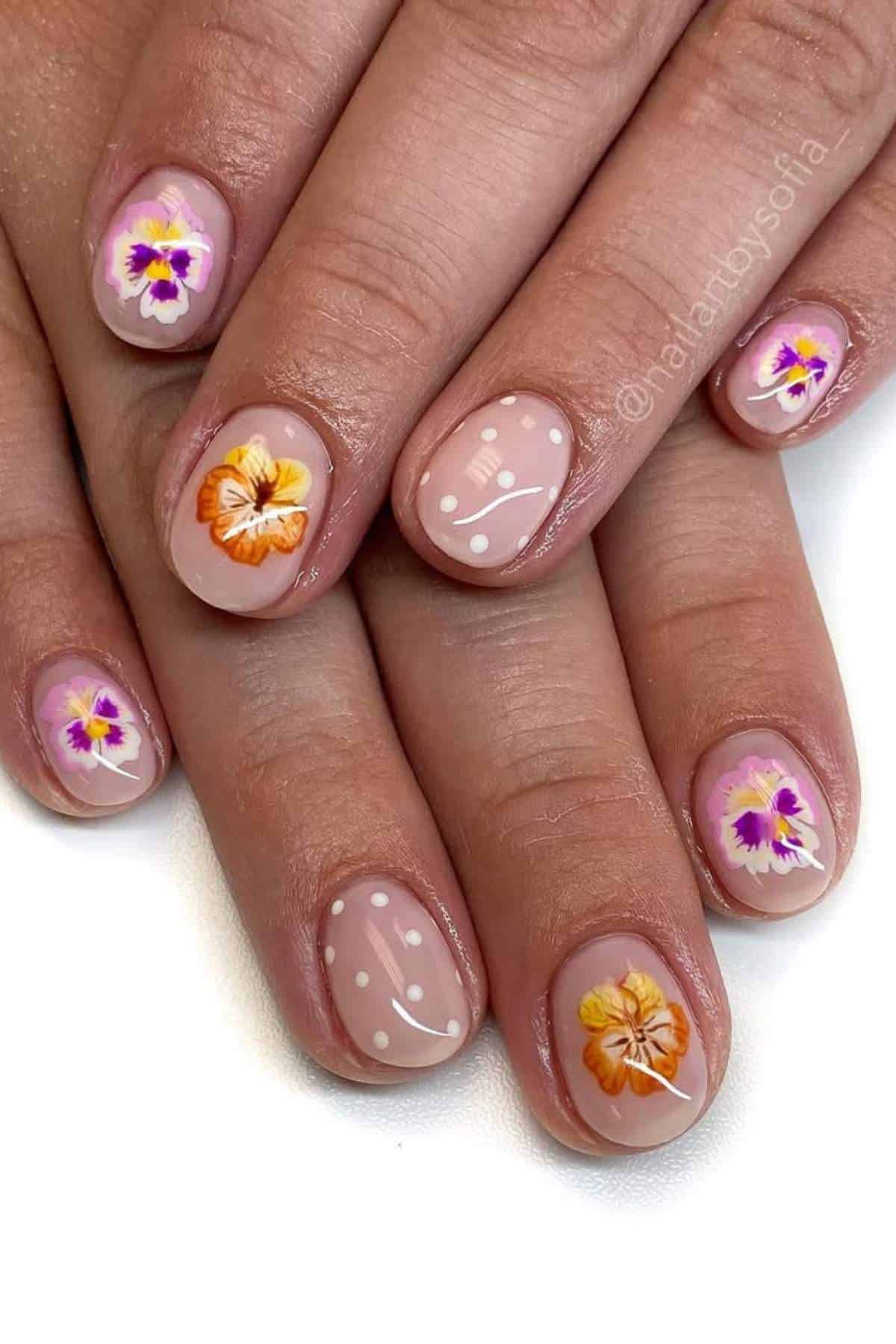 Hands with manicured nails featuring hand-painted pansy flowers in purple, orange, and yellow, and white polka dot designs on a nude base.