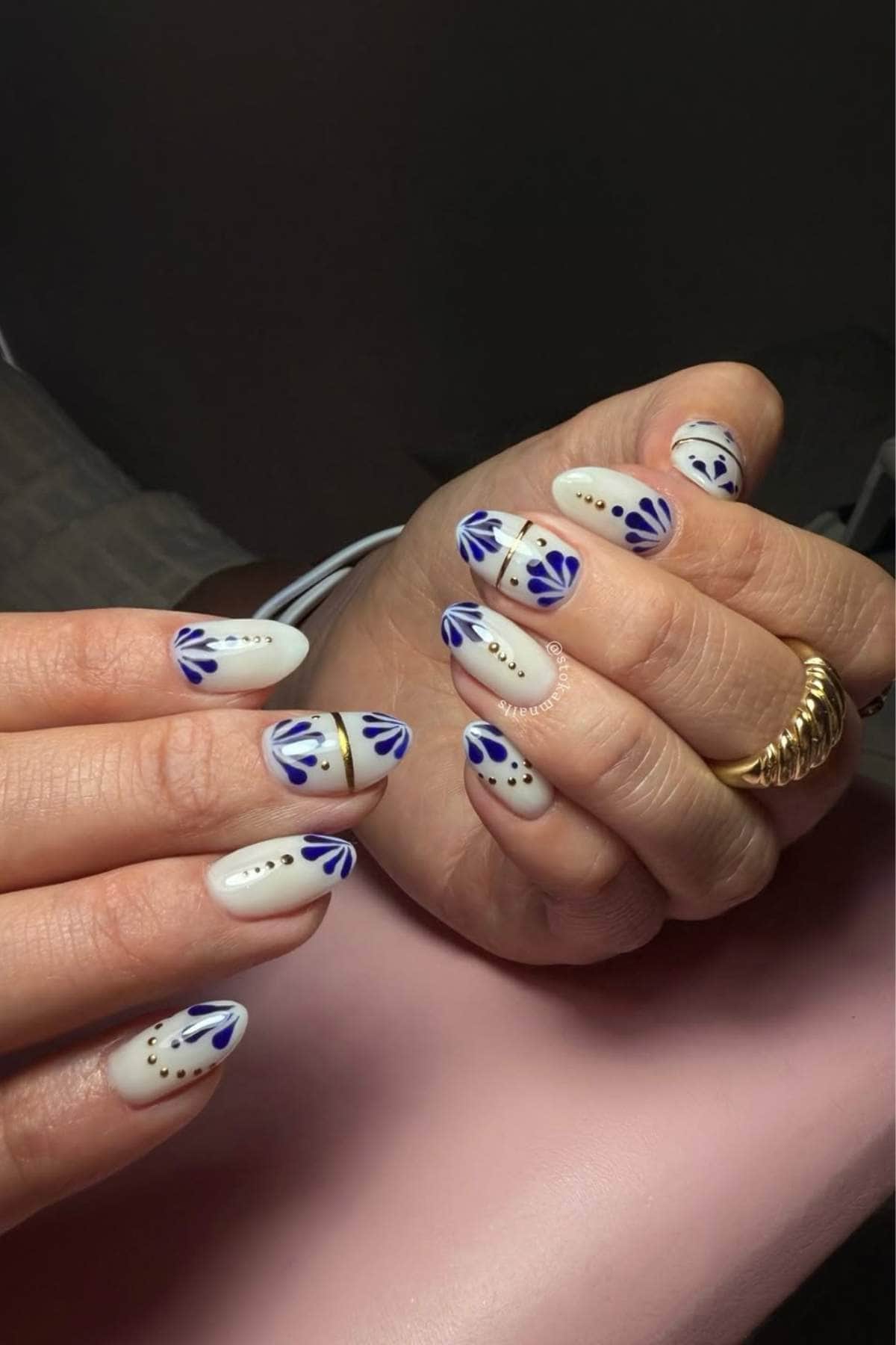 Hands with almond-shaped nails painted white, decorated with blue petal designs, gold lines, and small gold dots. The person wears gold rings.