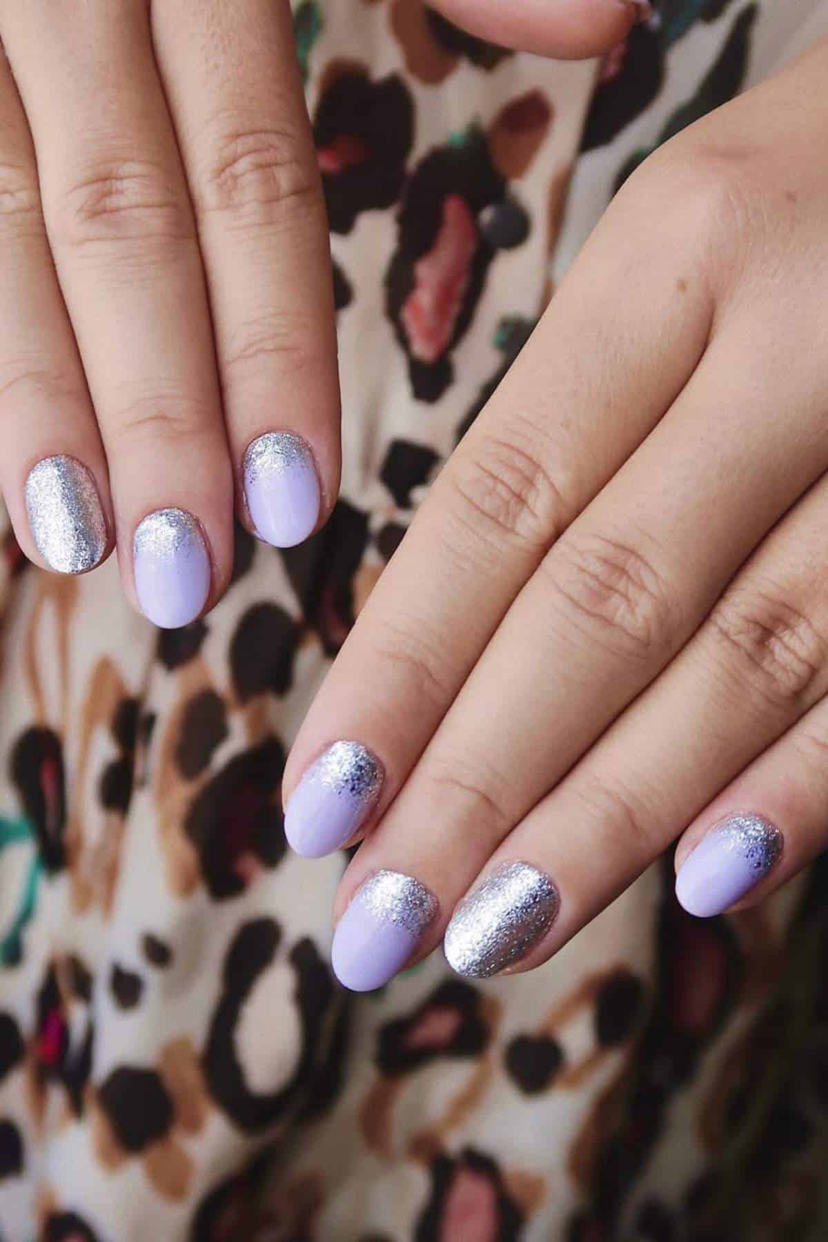 A close-up of hands with nails painted in light purple and silver glitter polish, against a background with a leopard print pattern.