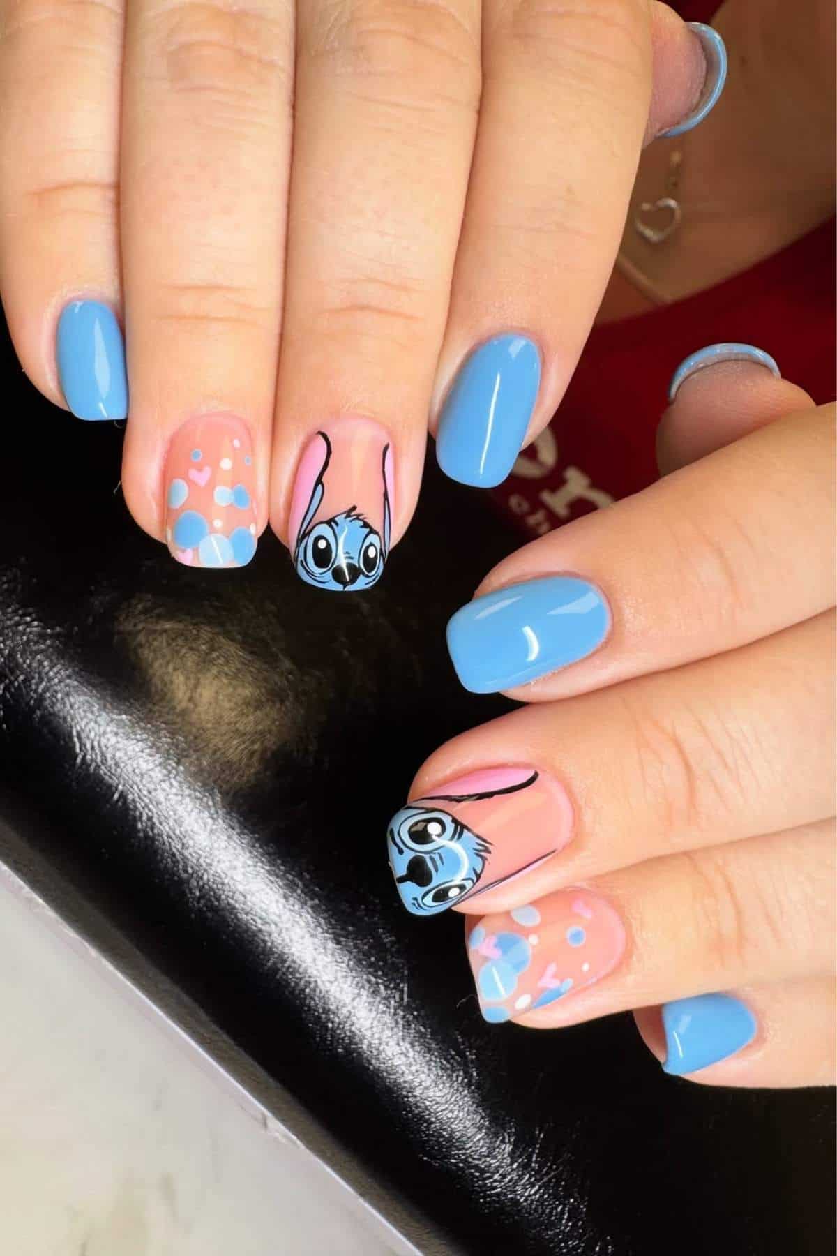 Hands with manicured nails featuring blue polish, pink accents, floral designs, and cartoon animal faces painted on two nails.
