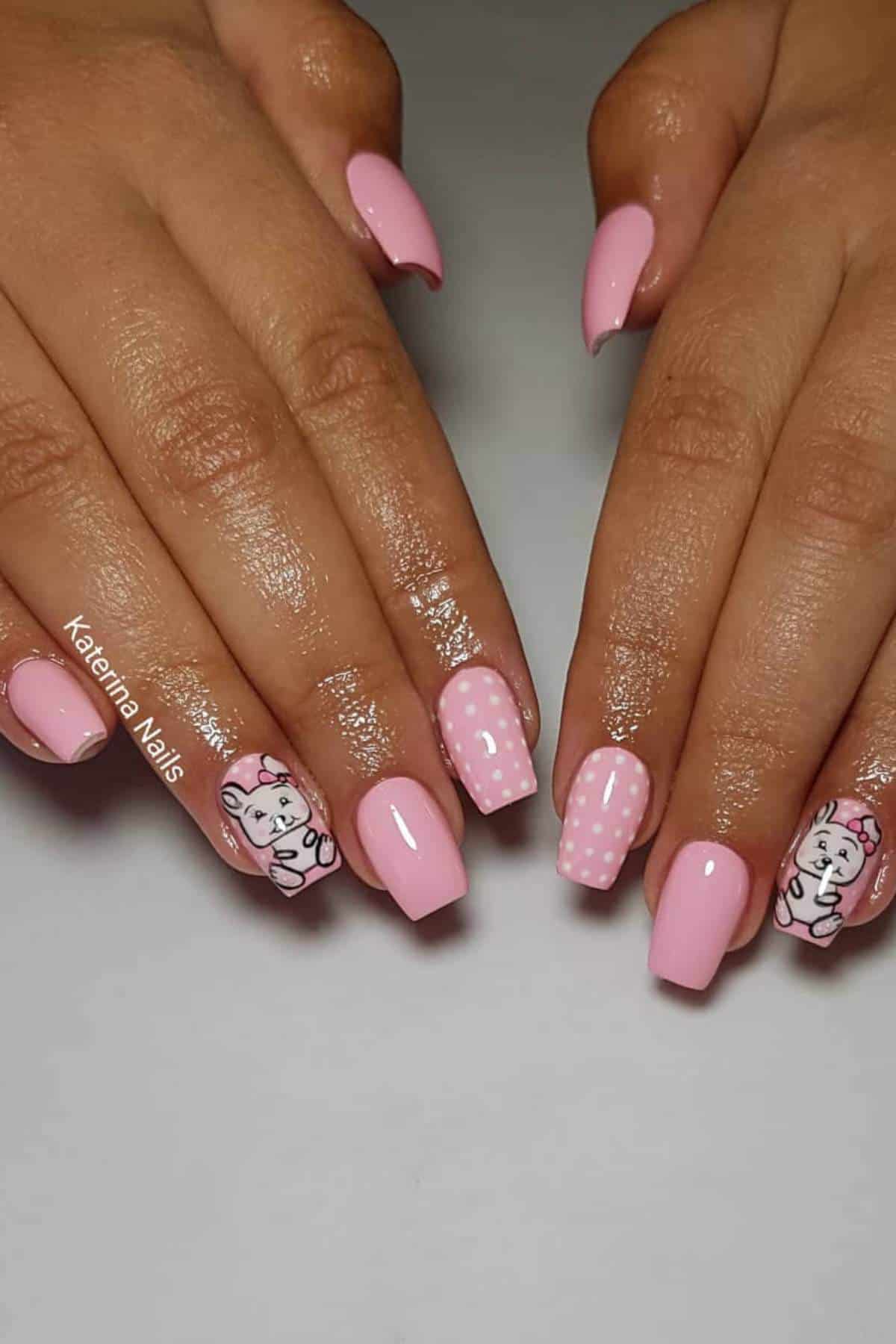 Hands with medium-length square nails painted pink, featuring cartoon cat faces and white polka dot designs on the ring and middle fingers.