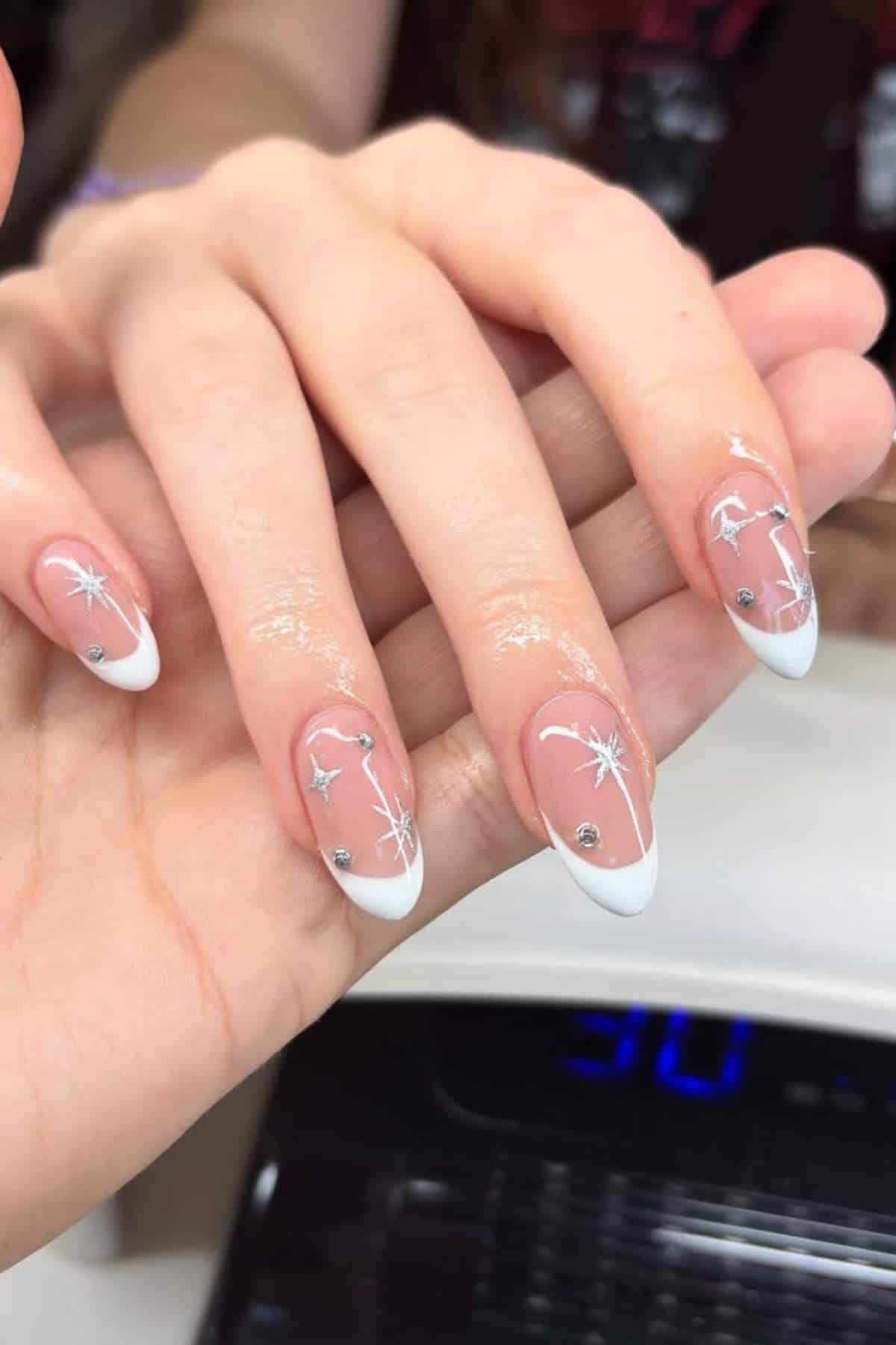 A hand with almond-shaped nails featuring a white French tip design, silver star-shaped decorations, and delicate white line accents.