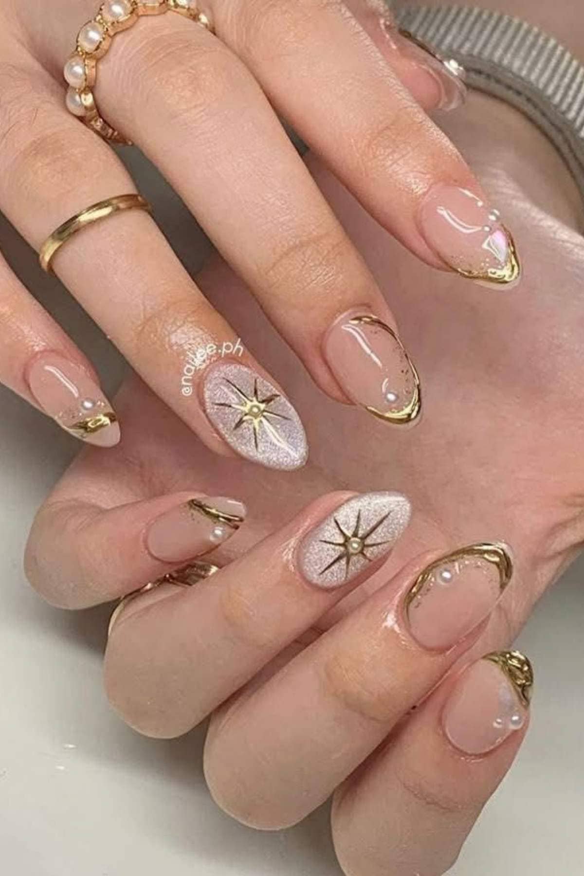 Close-up of hands with almond-shaped nails featuring nude polish, gold tips, pearl accents, and star designs on a glittery background.
