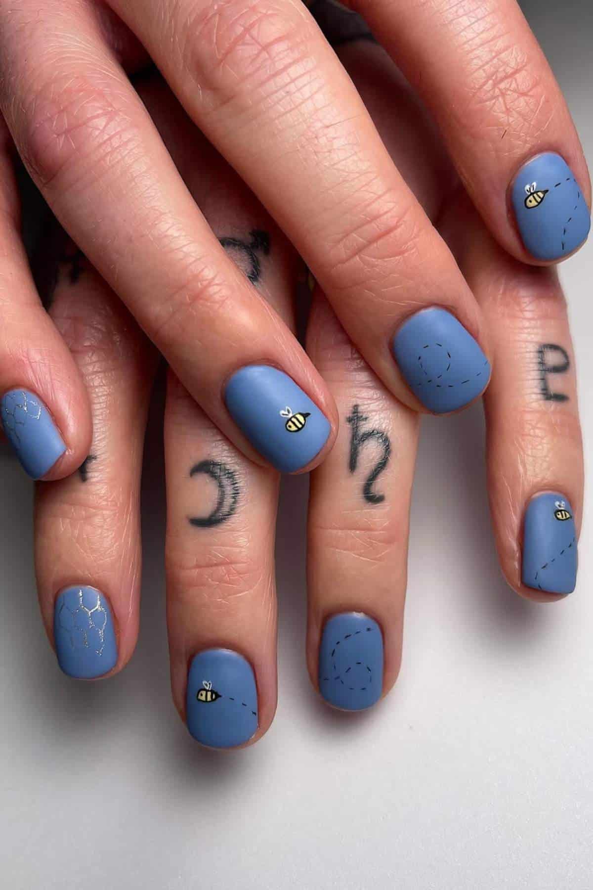 Hands with blue nail polish, adorned with bee designs and dotted lines, rest over fingers featuring black-ink tattoos.
