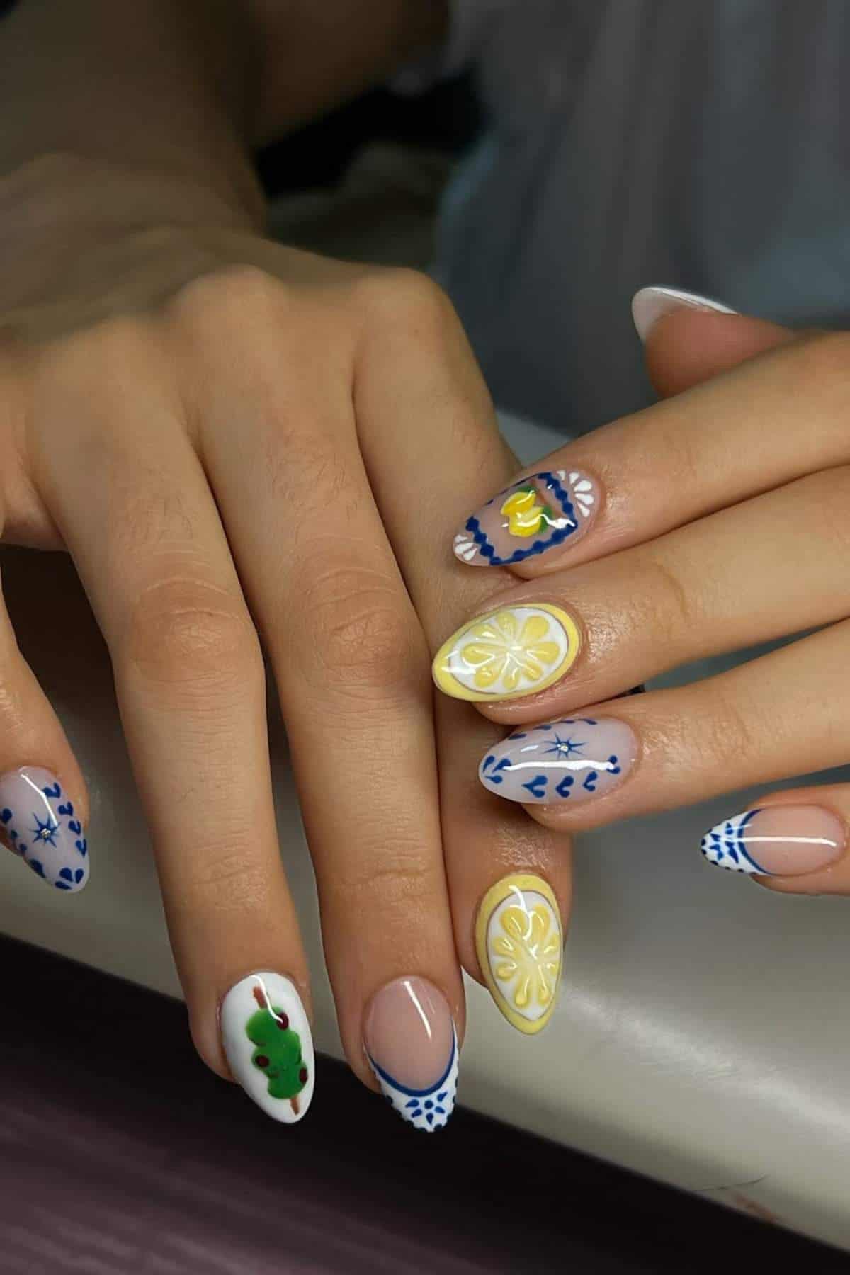 A hand with long nails featuring intricate nail art, including blue patterns, yellow lemons, and green shapes on a neutral base.