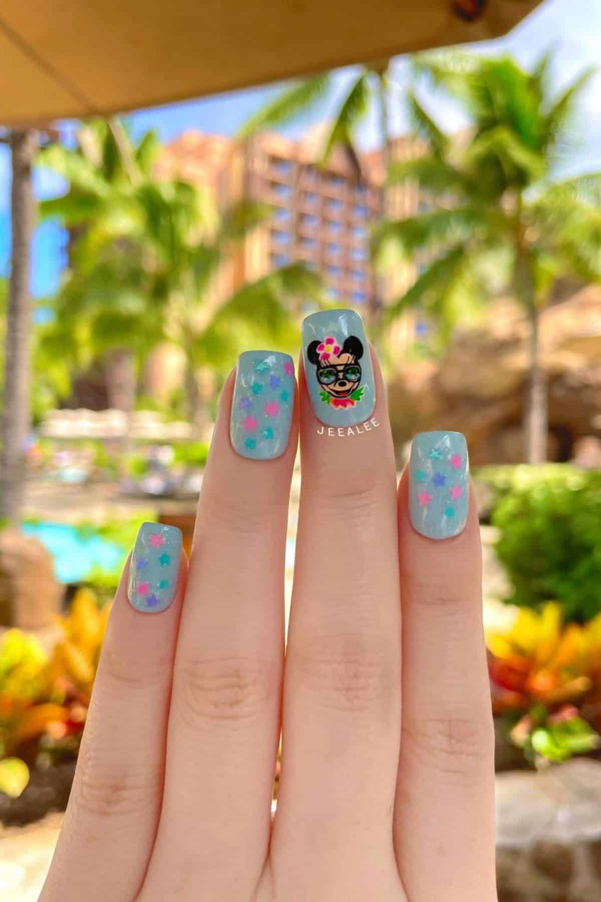 A hand with long, blue-painted nails featuring pink and light blue star designs, and a Mickey and Minnie Mouse decal, is held up outdoors with palm trees and a resort in the background.
