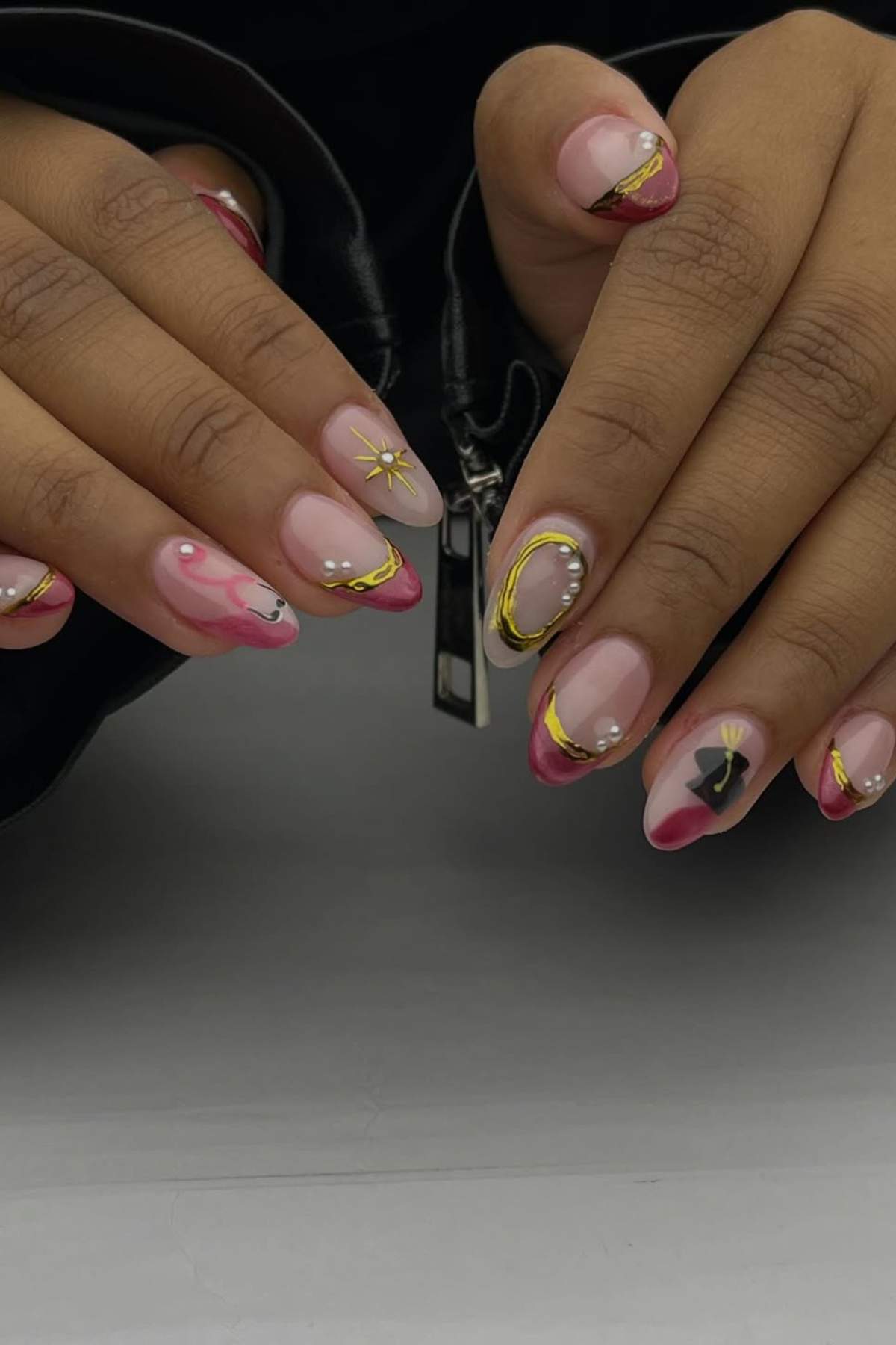 Hands with long almond-shaped nails featuring pink tips, gold and black abstract designs, pearls, and small star accents, against a neutral background.
