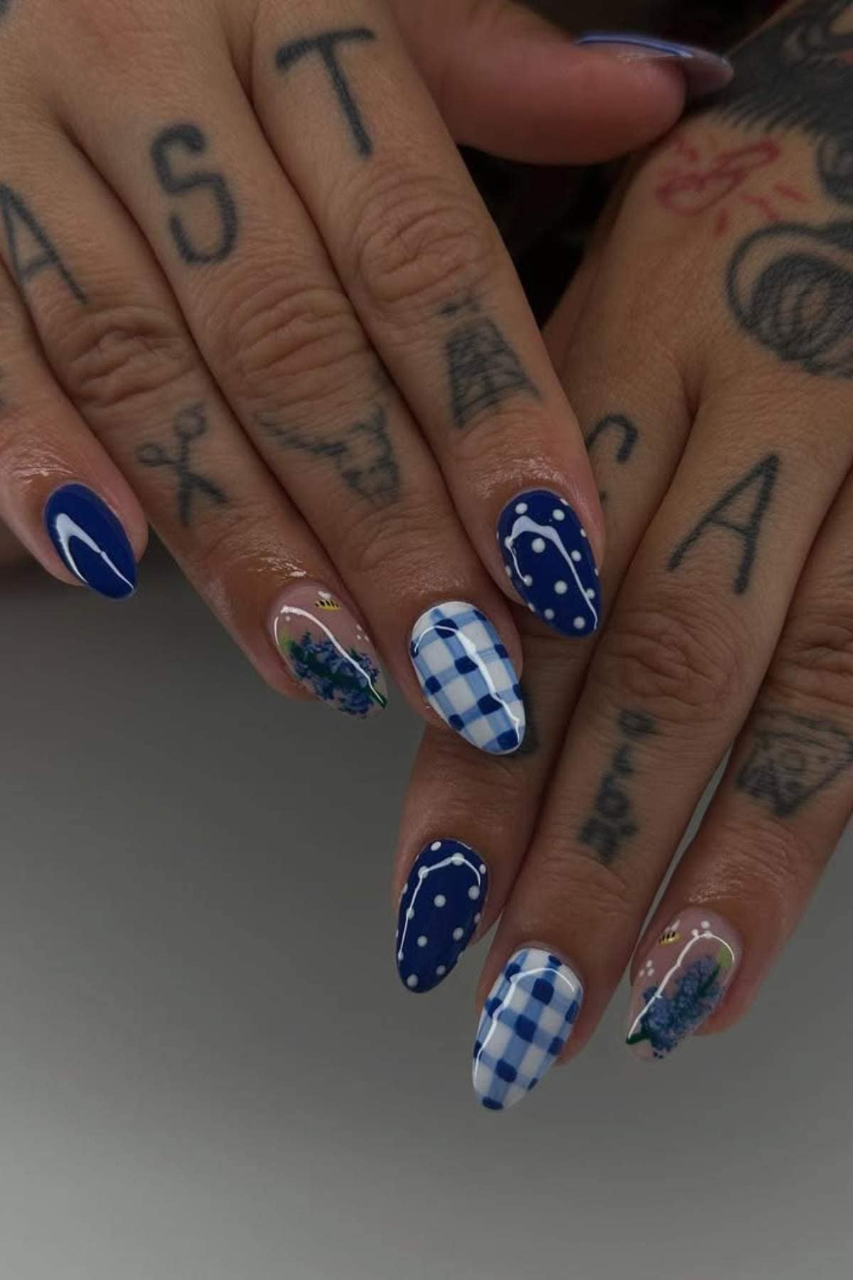 Hands with tattoos showing manicured nails featuring blue and white designs, including plaid, polka dots, floral patterns, and solid blue with a white crescent.