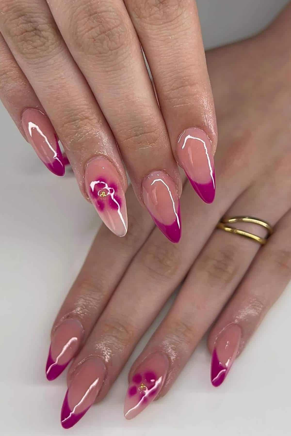 Hands with almond-shaped nails, featuring pink French tips and two accent nails with pink floral designs and gold details. The person wears two gold rings on one finger.
