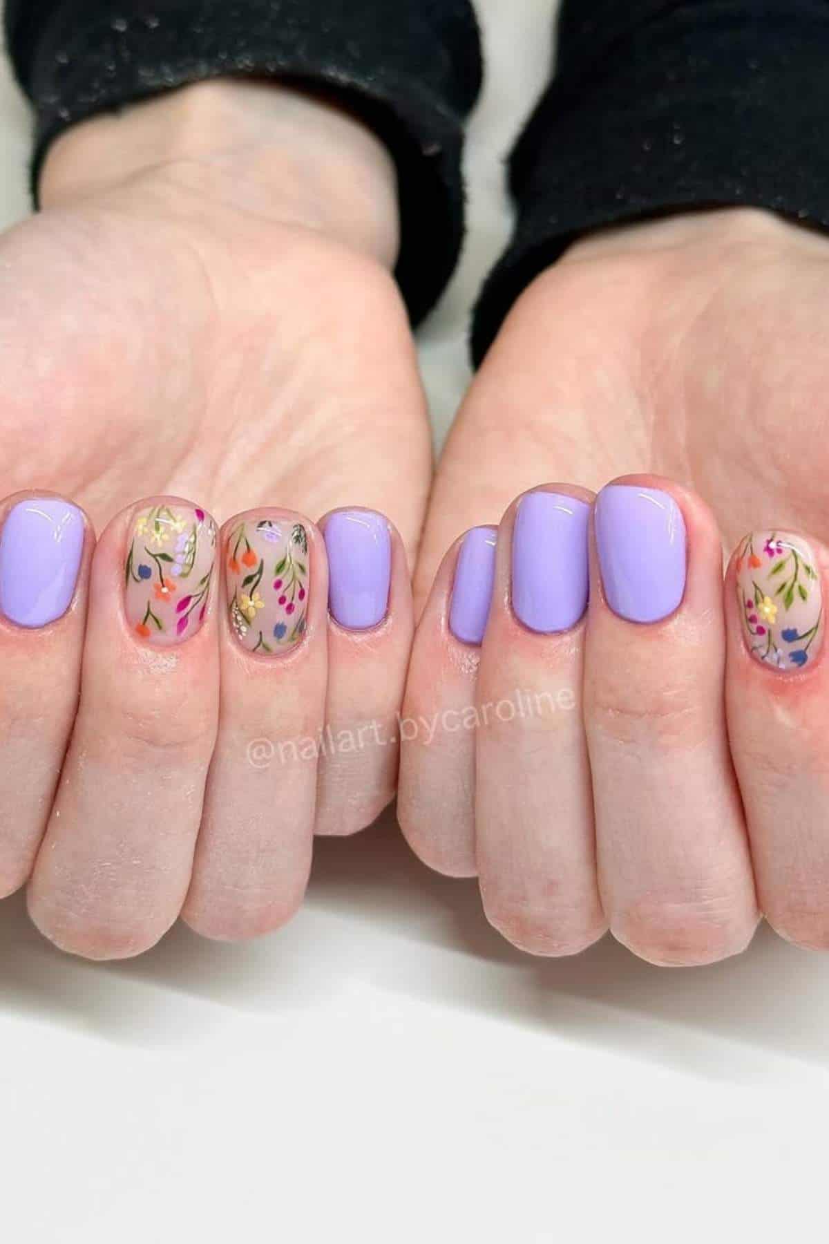 Hands with light purple nail polish; ring and middle fingers feature a clear base with colorful floral designs.
